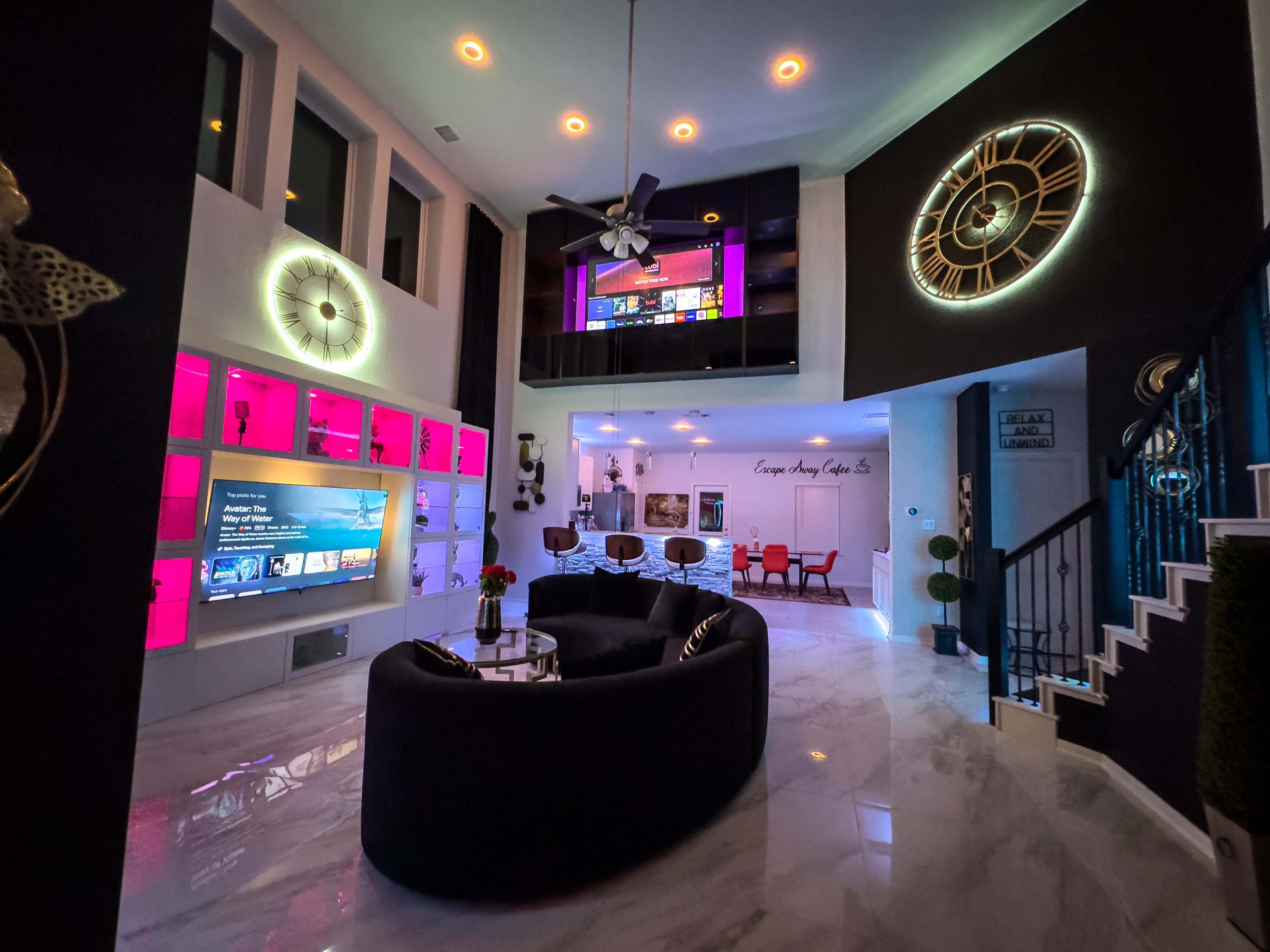 A modern living area featuring a circular sofa, large television, decorative wall clocks, and vibrant lighting fixtures.