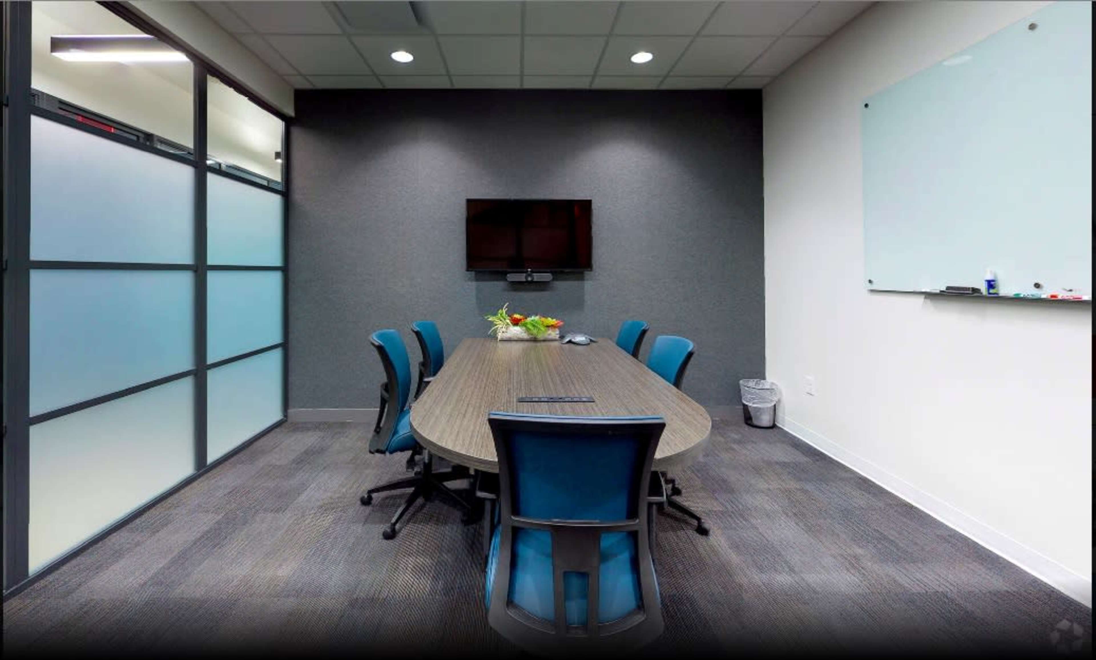 The image shows a modern conference room with a long table, six blue chairs, a wall-mounted television, and a whiteboard.