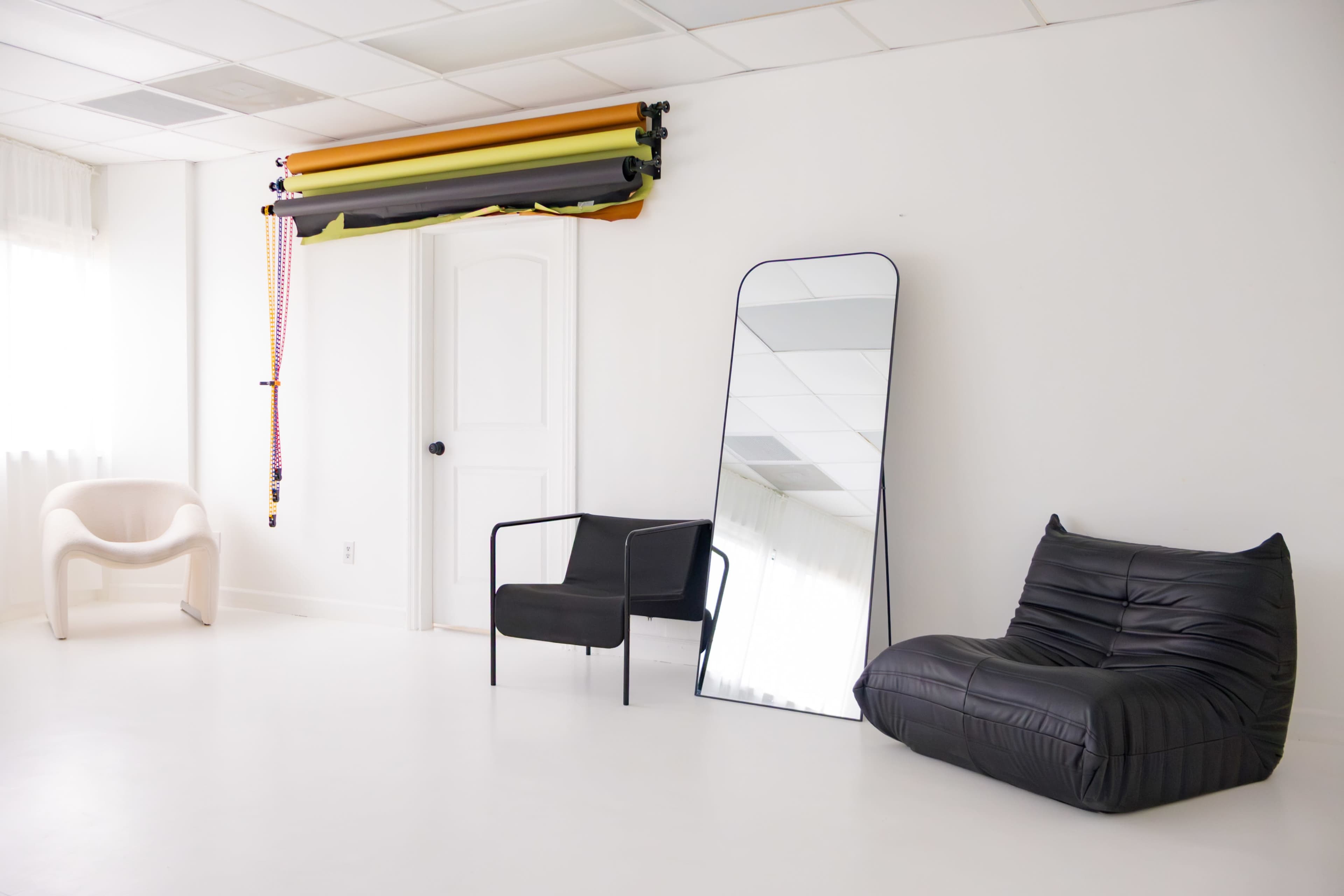The image features a minimalist studio space with a black lounge chair, a white armchair, a full-length mirror, and a roll of backdrop paper hanging on the wall.