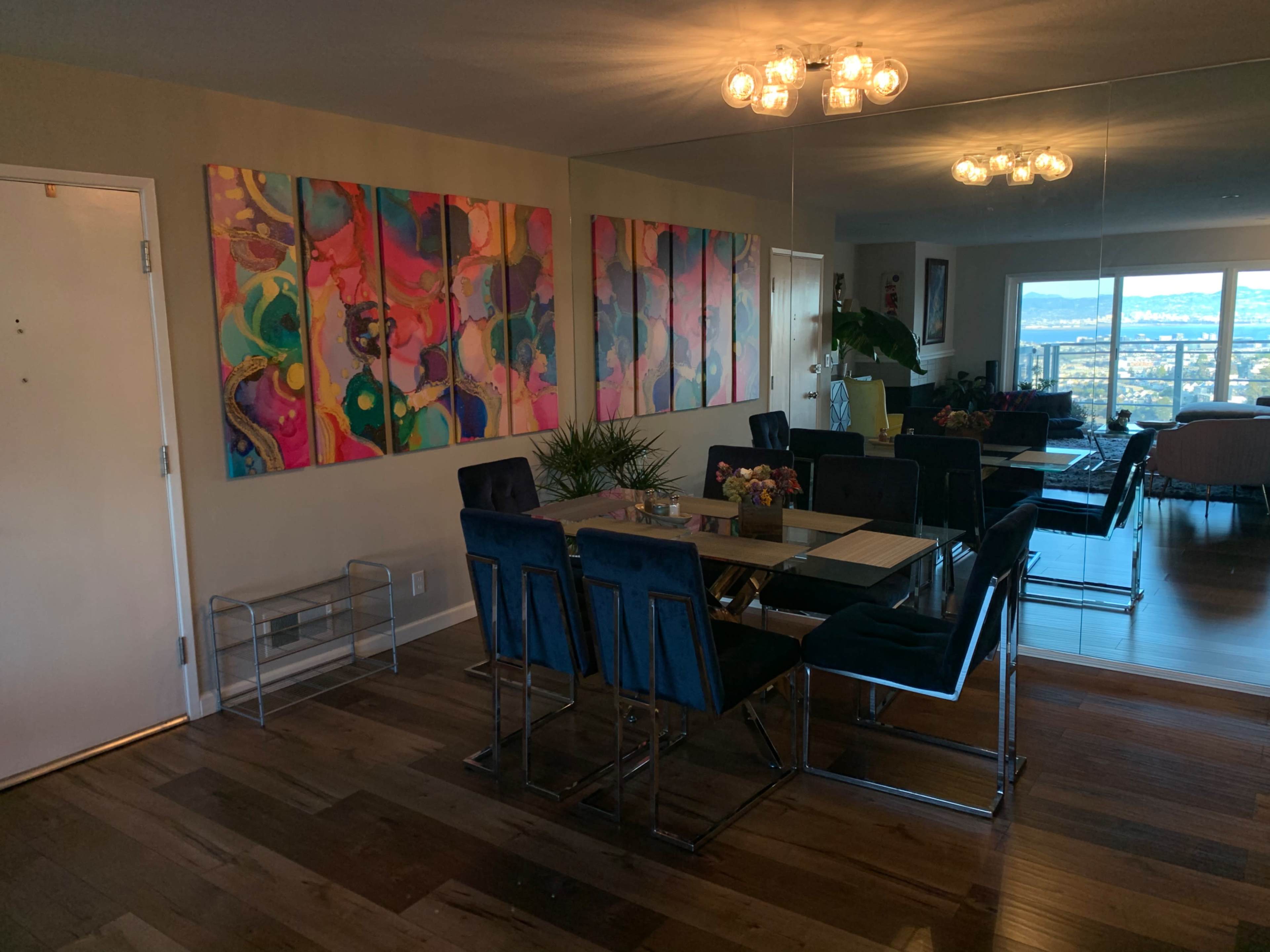 A dining area featuring a large table surrounded by chairs, with a colorful abstract artwork displayed on the wall and large windows providing a view of the city.