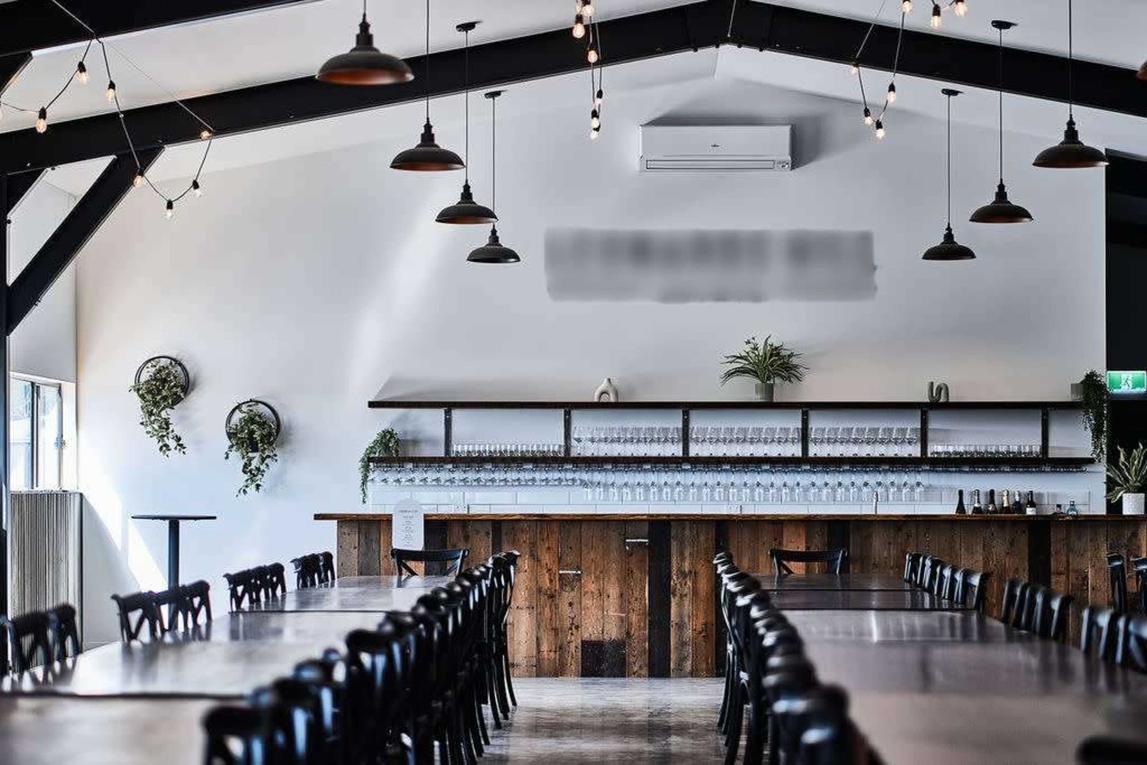 A modern dining space featuring long tables, a wooden bar with shelves, and pendant lighting against a high, slanted ceiling.
