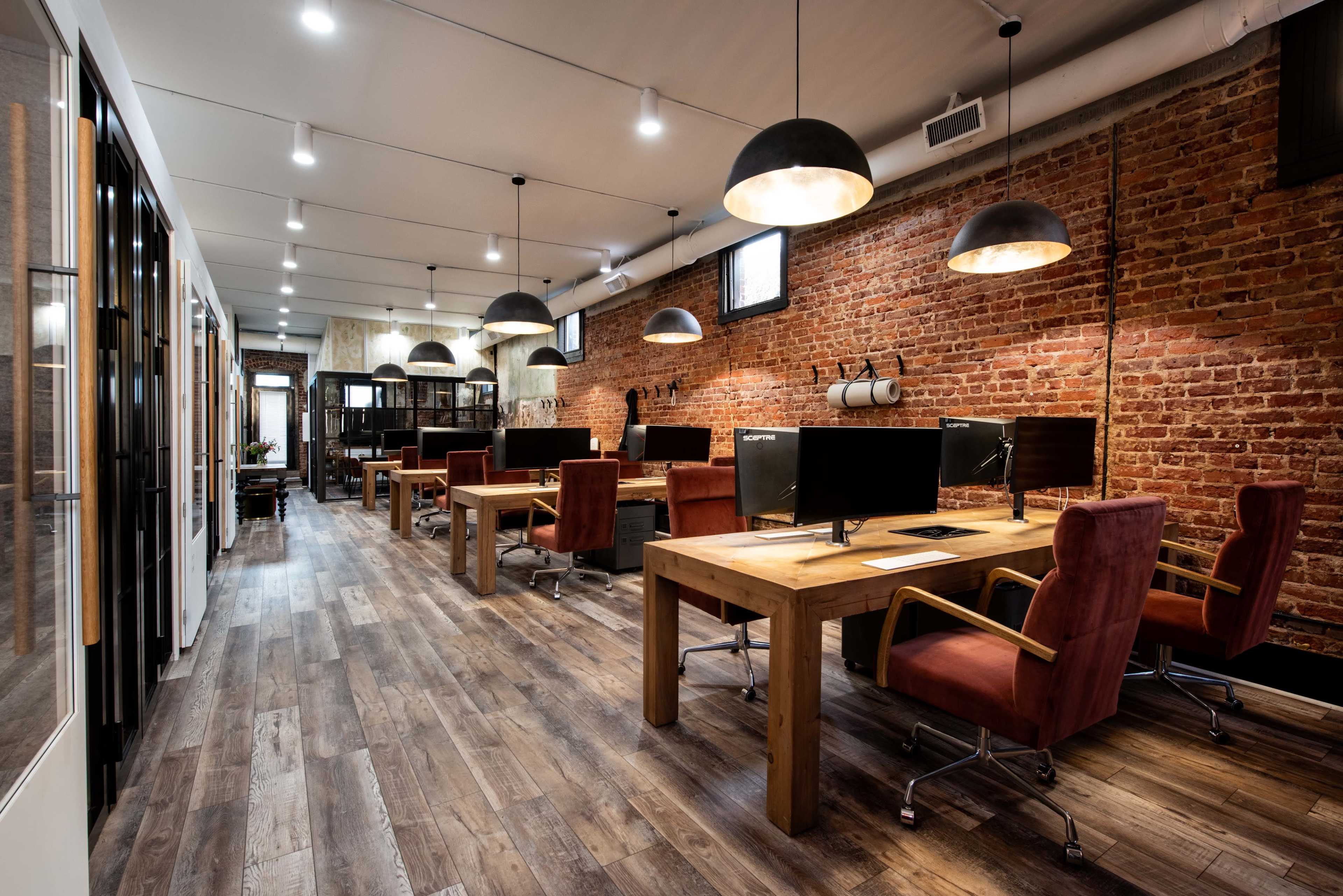 A modern office space featuring several wooden desks with computers, accentuated by exposed brick walls and pendant lighting.