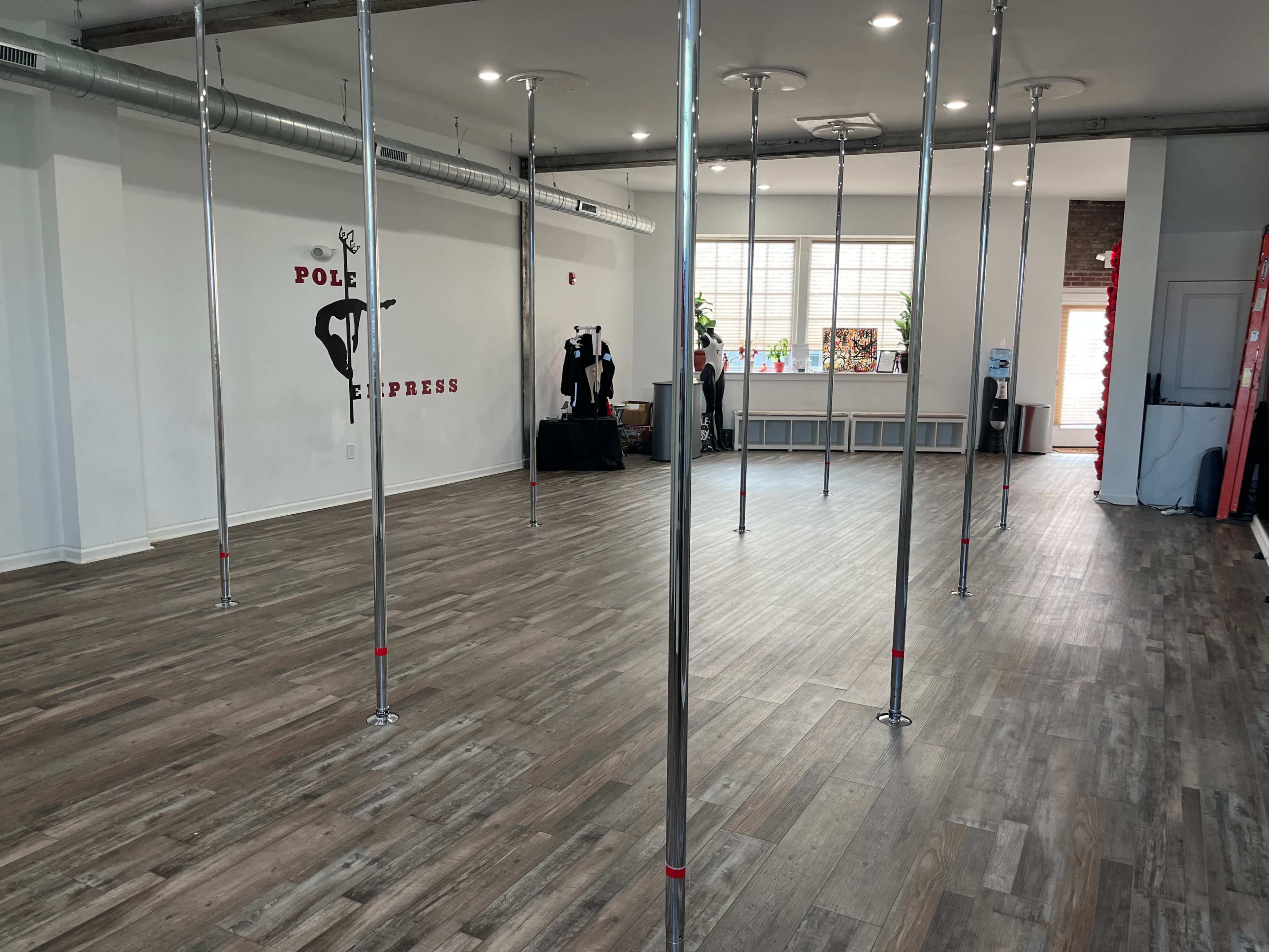 The image shows an empty dance studio with several chrome poles arranged on a wooden floor and a backdrop featuring the words "Pole Express" on the wall.