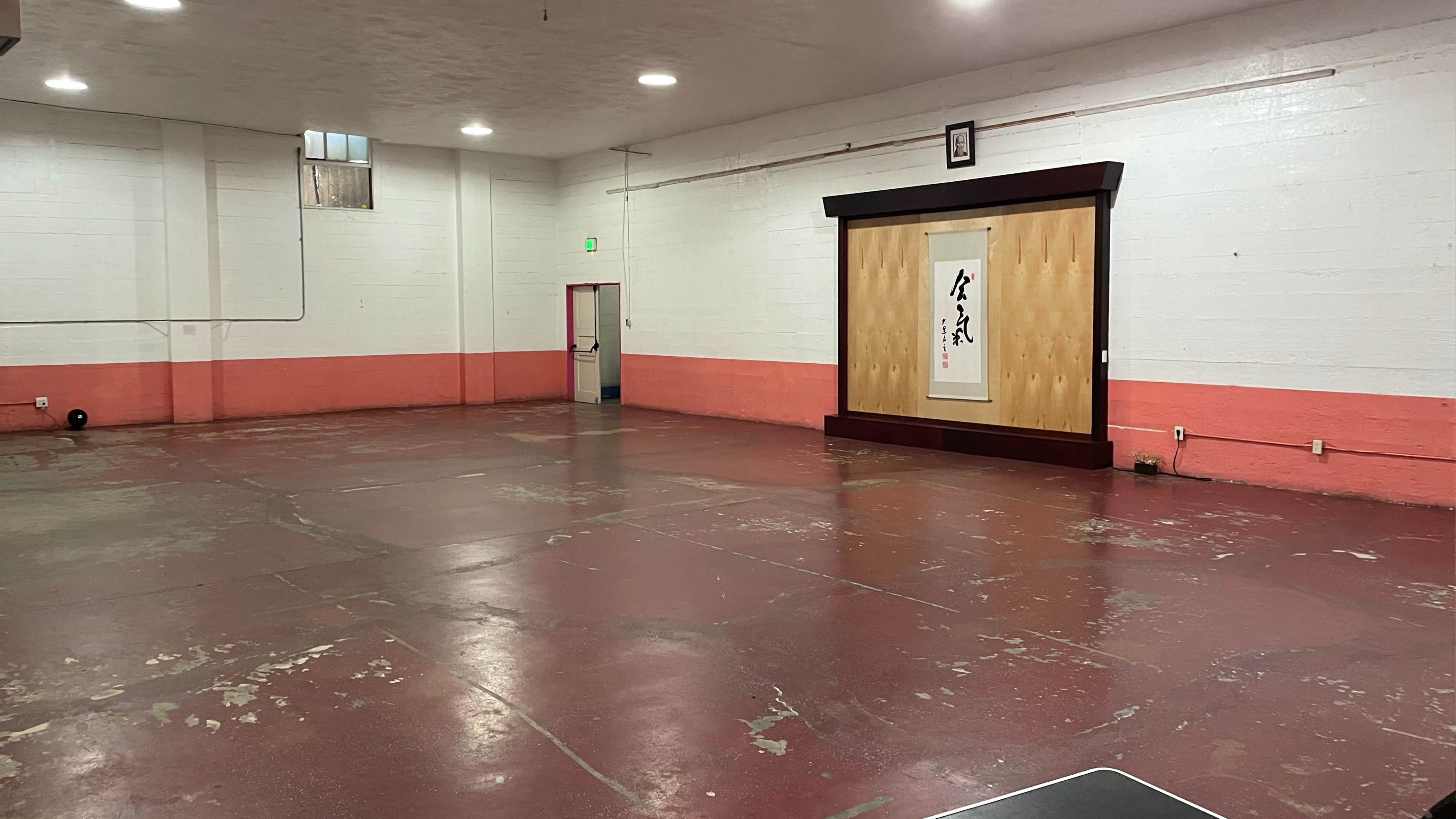 The image shows an empty, industrial-style room with concrete walls, a red-painted lower section, and a wooden wall feature displaying Asian calligraphy.