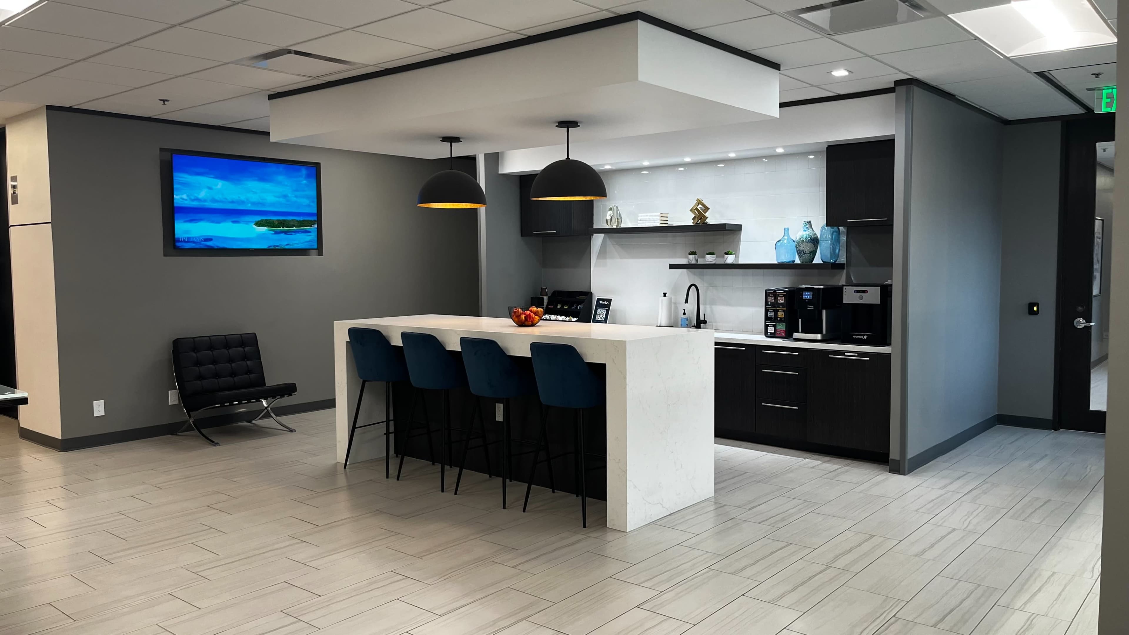 A modern office kitchen area featuring a large white island with bar stools, a coffee station, and a wall-mounted television displaying a landscape.