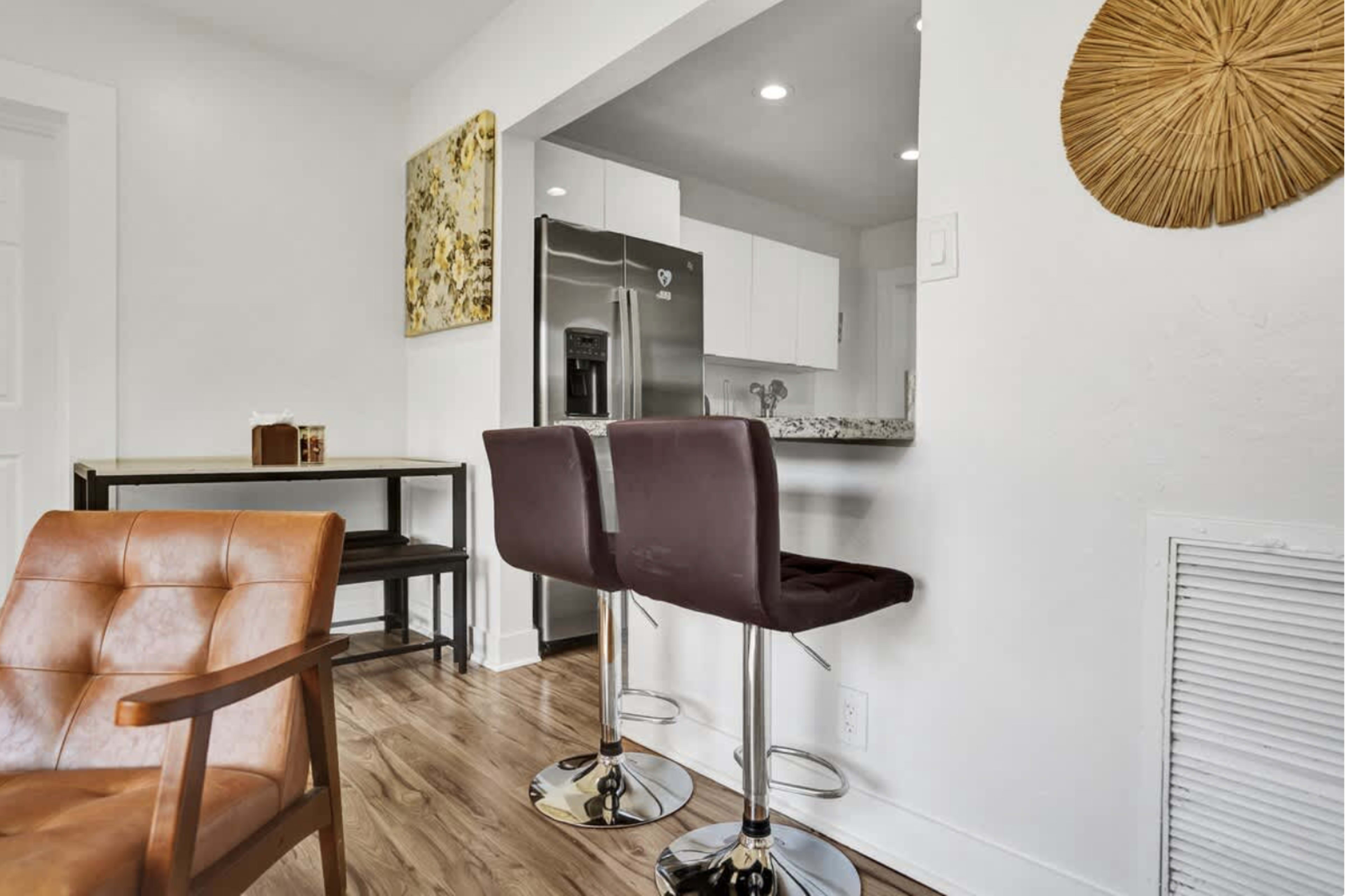 The image shows a modern interior featuring two bar stools at a kitchen counter, a brown leather armchair, and a wooden table against a light-colored wall.