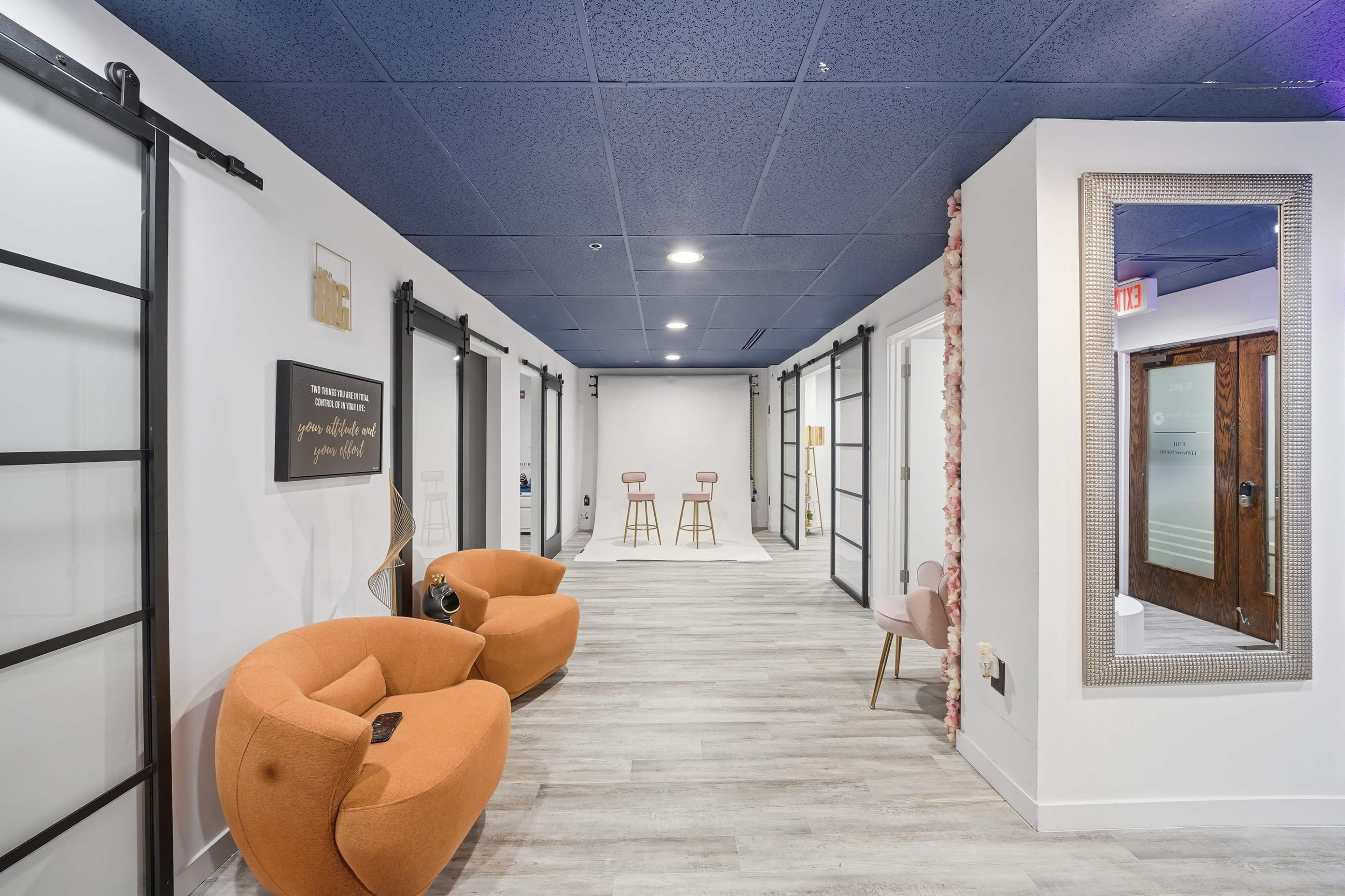 A well-lit hallway features a pair of orange chairs, multiple closed doors on either side, and a mirror with a floral border at one end.
