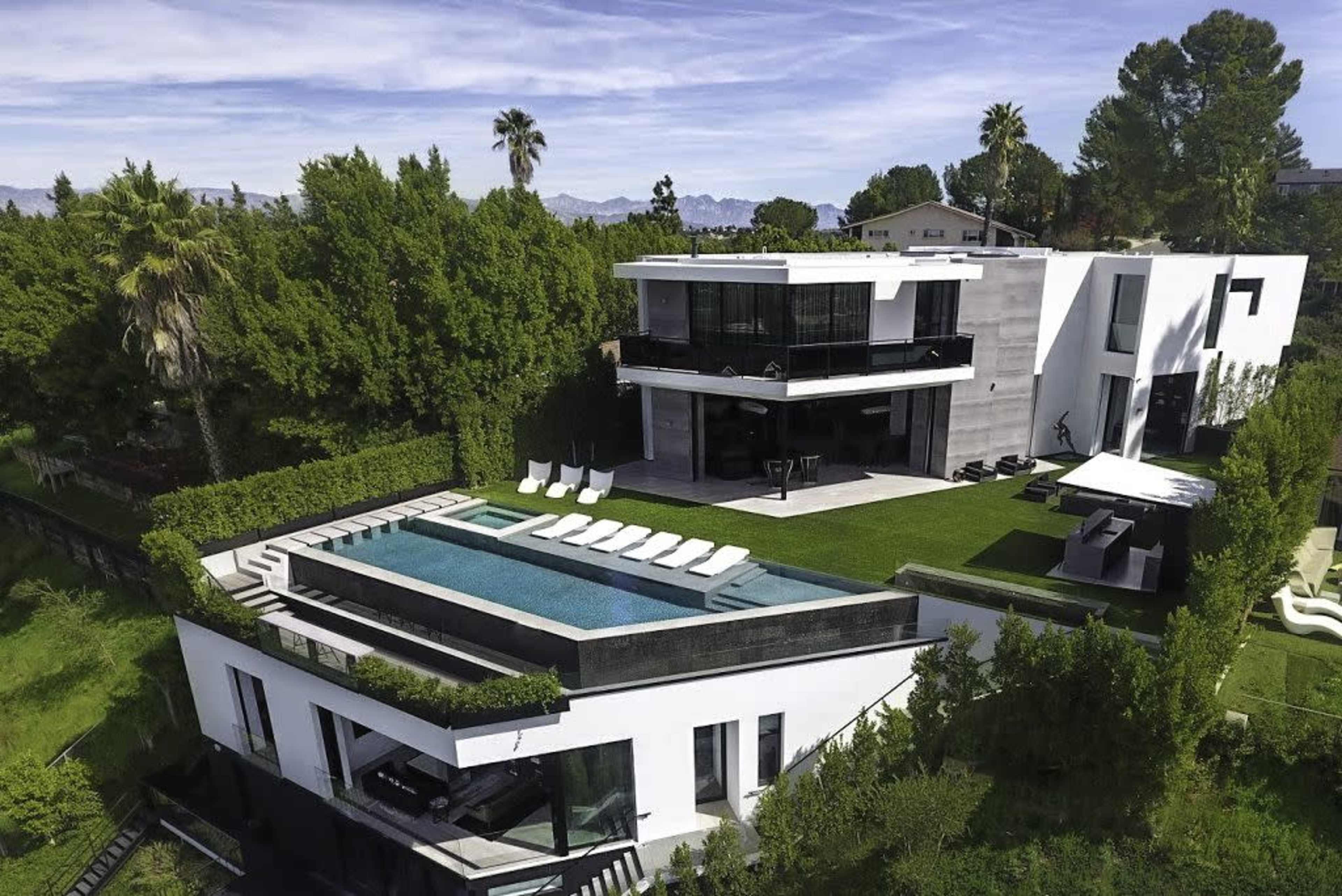 The image shows a modern multi-level house with a swimming pool, surrounded by greenery and palm trees.