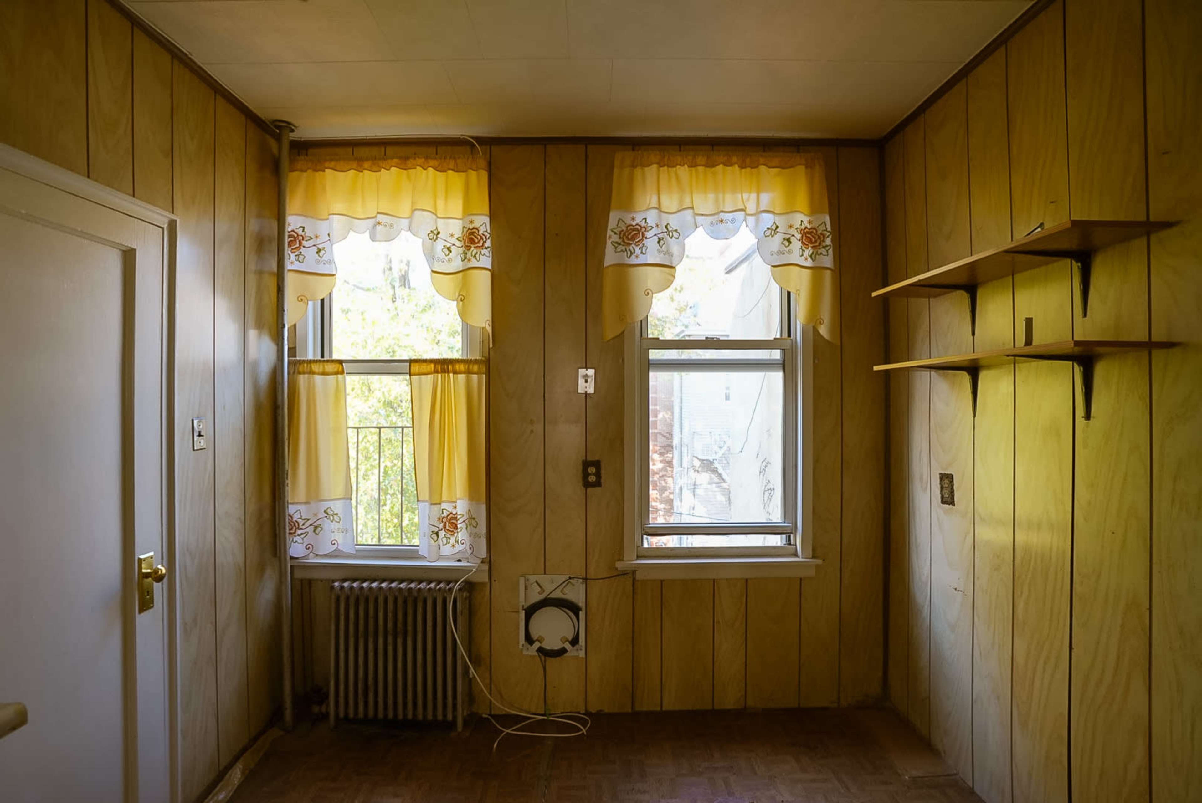The image shows a room with wooden paneling, two windows dressed with yellow curtains, and an empty shelf on the wall.