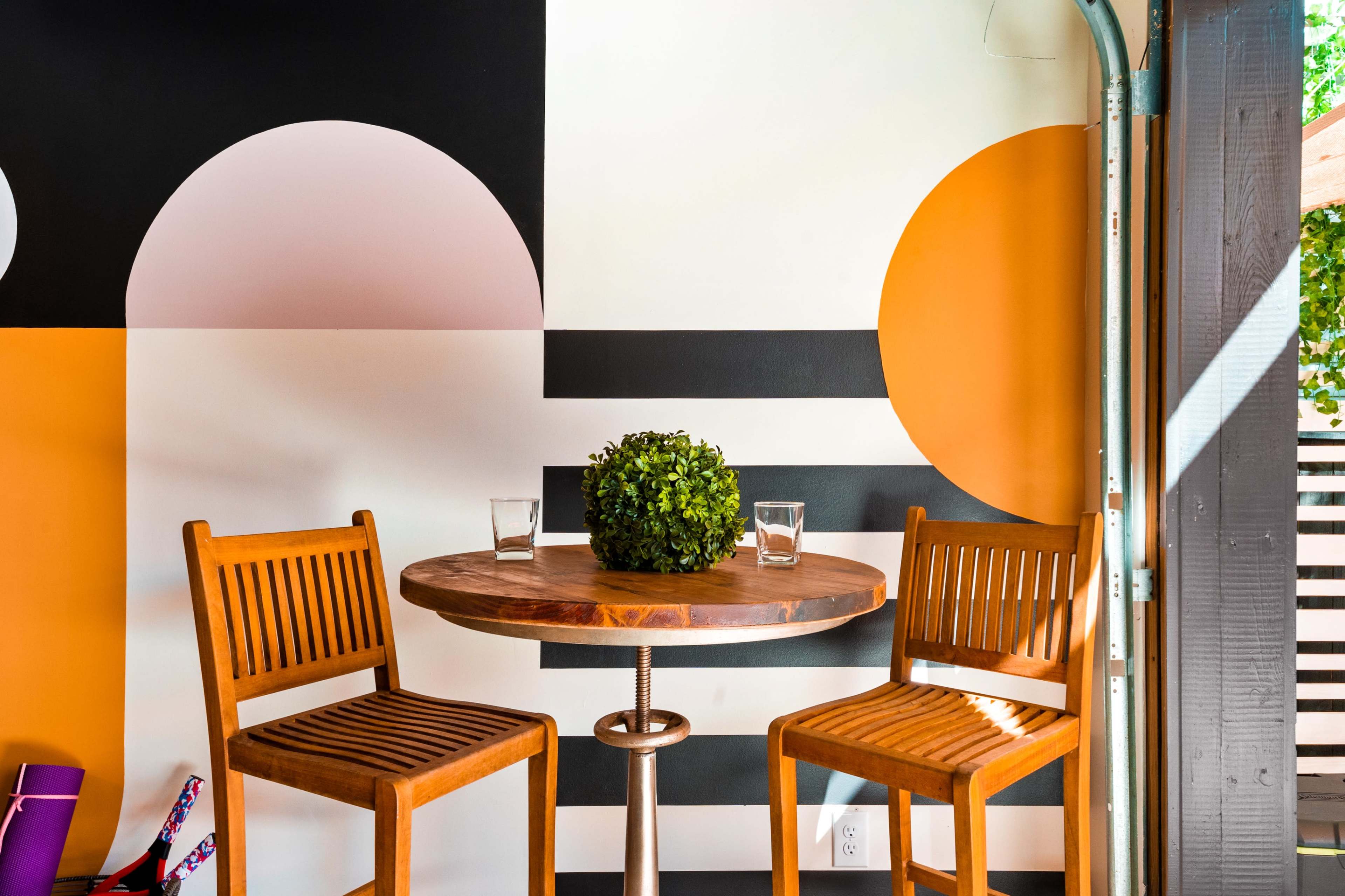 A round table with two wooden chairs is positioned against a wall featuring geometric shapes in orange, black, and cream colors, with a small plant centerpiece and two glasses on the table.