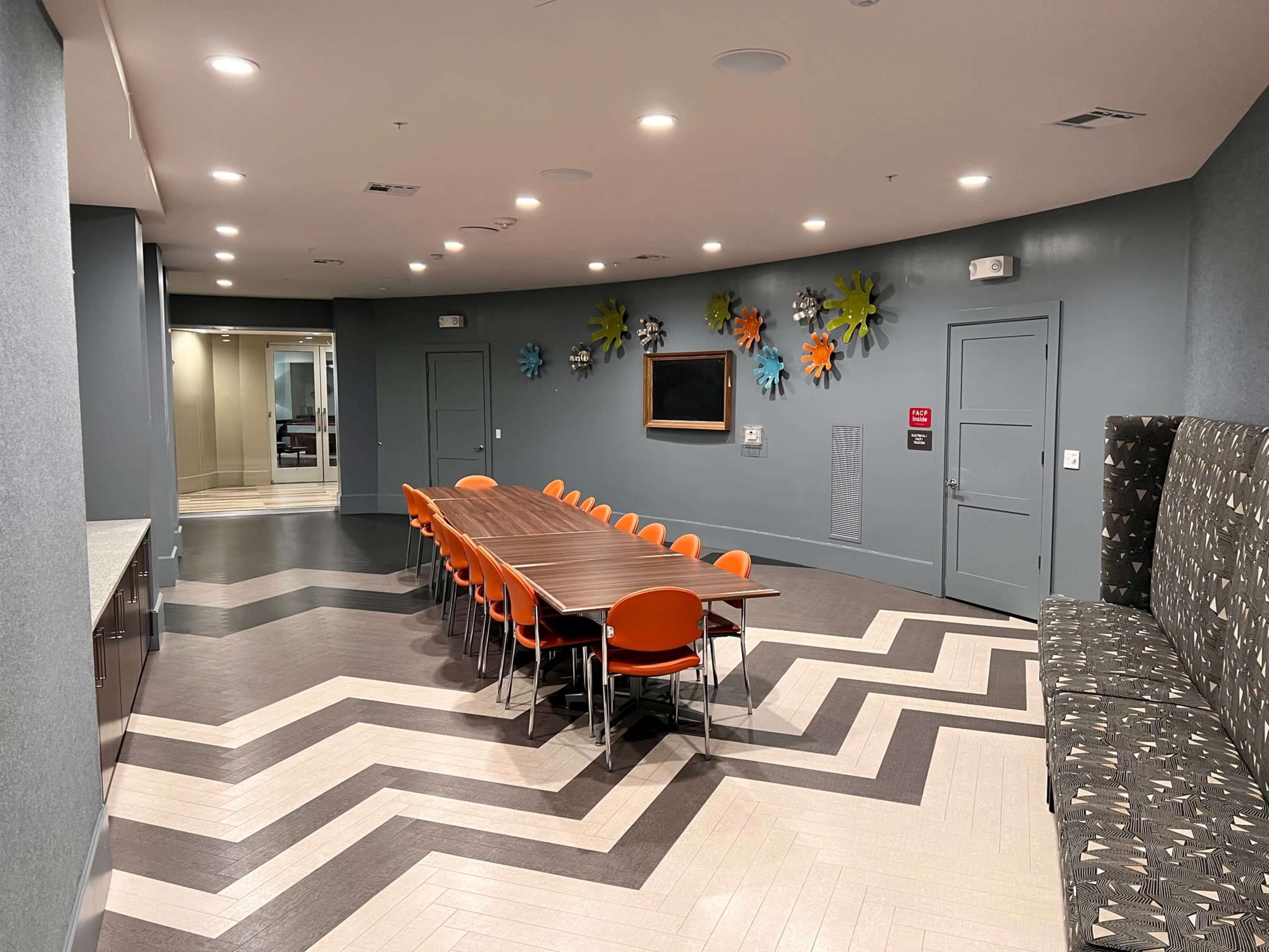A modern conference room with a long wooden table surrounded by orange chairs, geometric flooring, and decorative wall art.