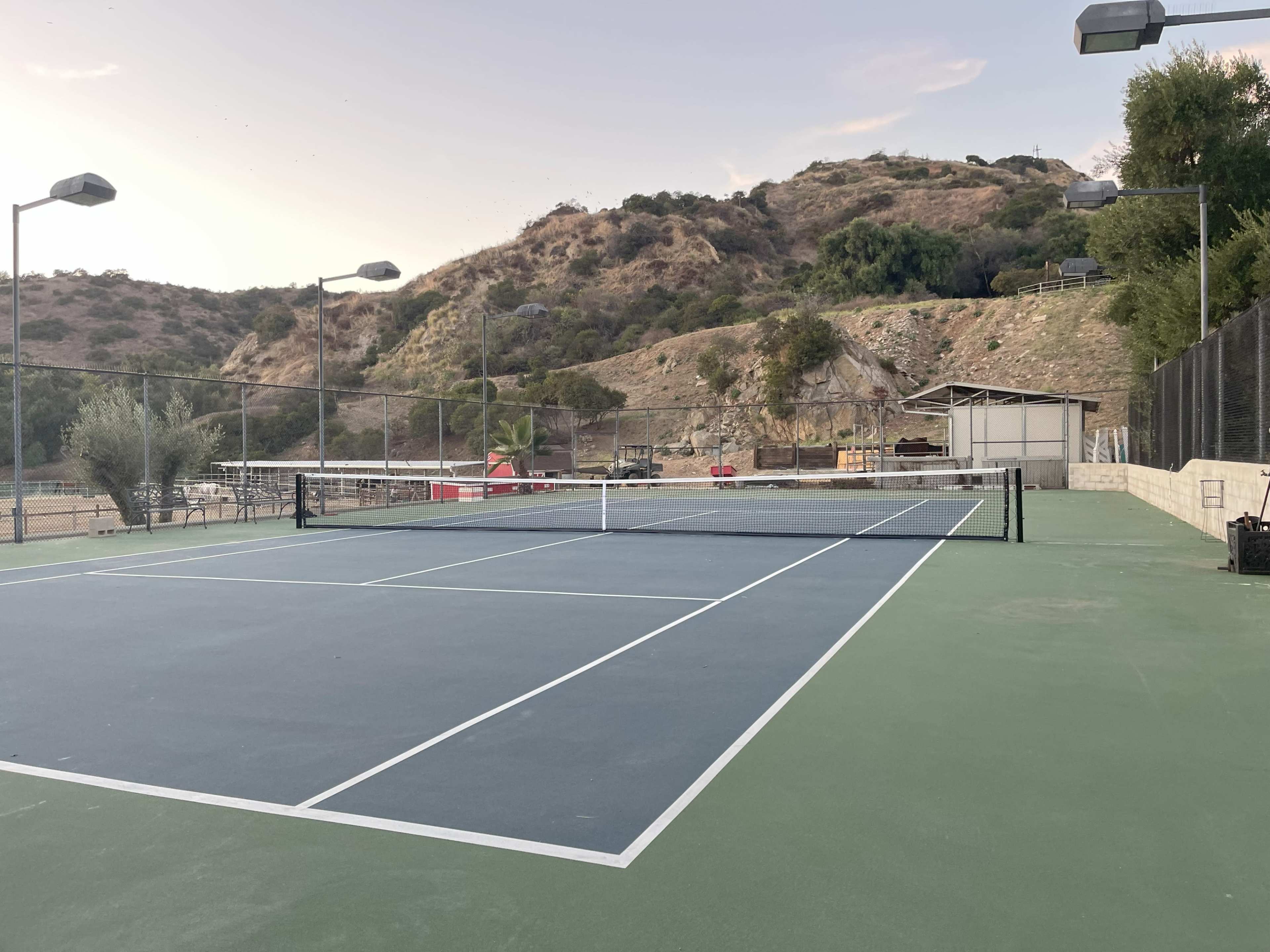 A tennis court with a green surface, situated in an outdoor setting with a hillside in the background and overhead lighting fixtures.
