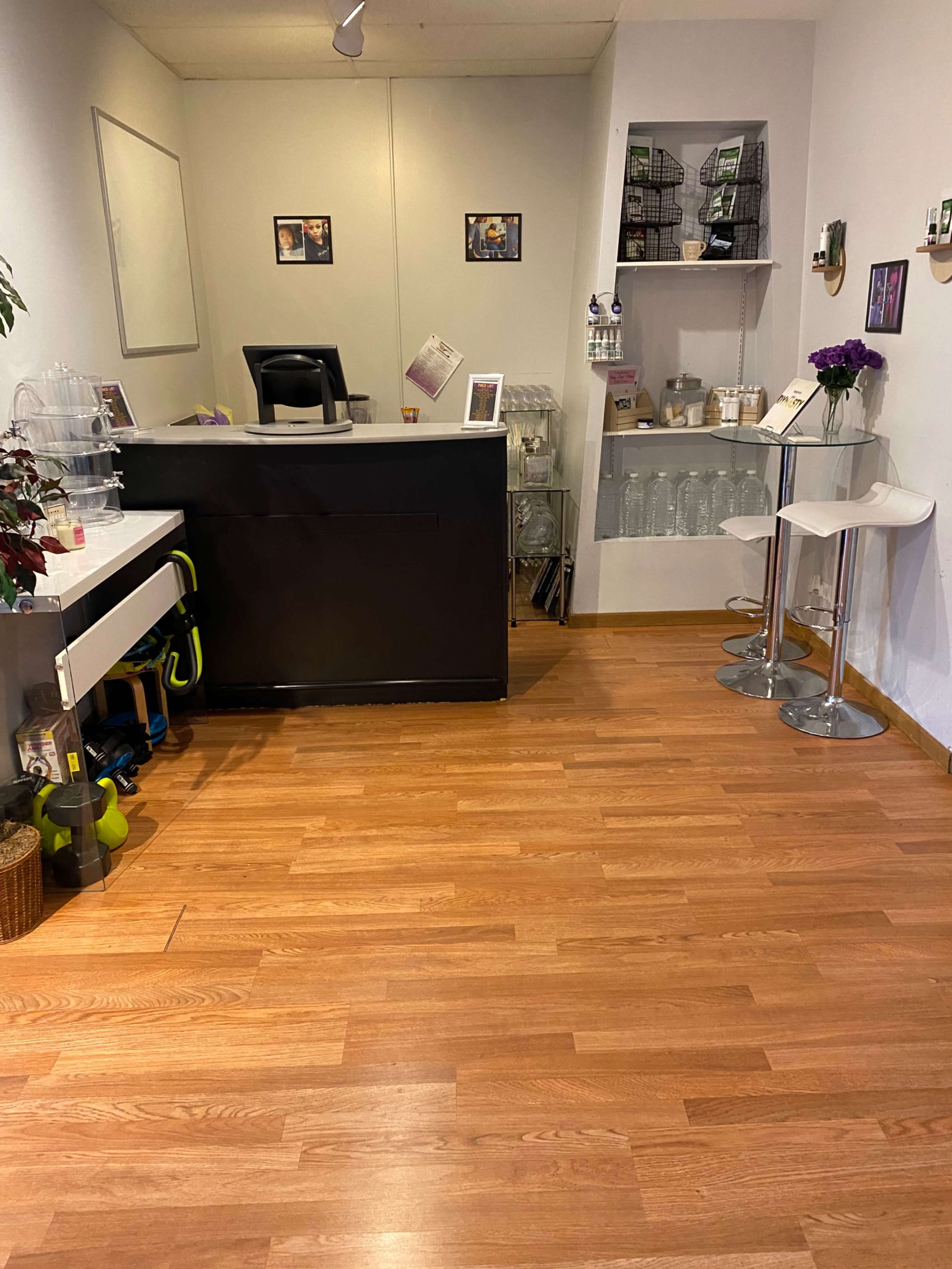 A small, modern reception area with a black front desk, white stools, a beverage station, and wooden flooring.
