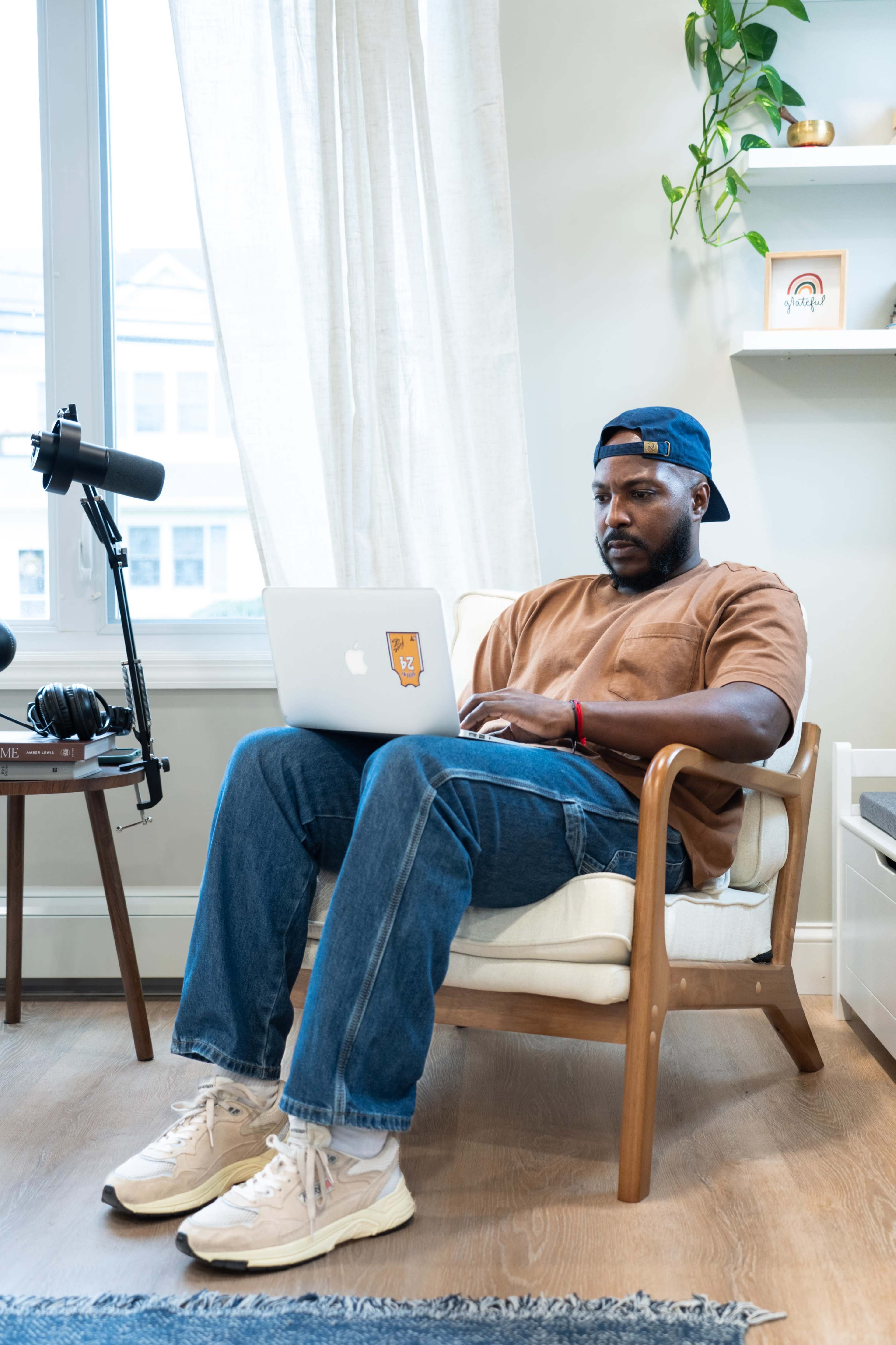 A man sits in a chair working on a laptop in a well-lit room with a microphone and headphones nearby.