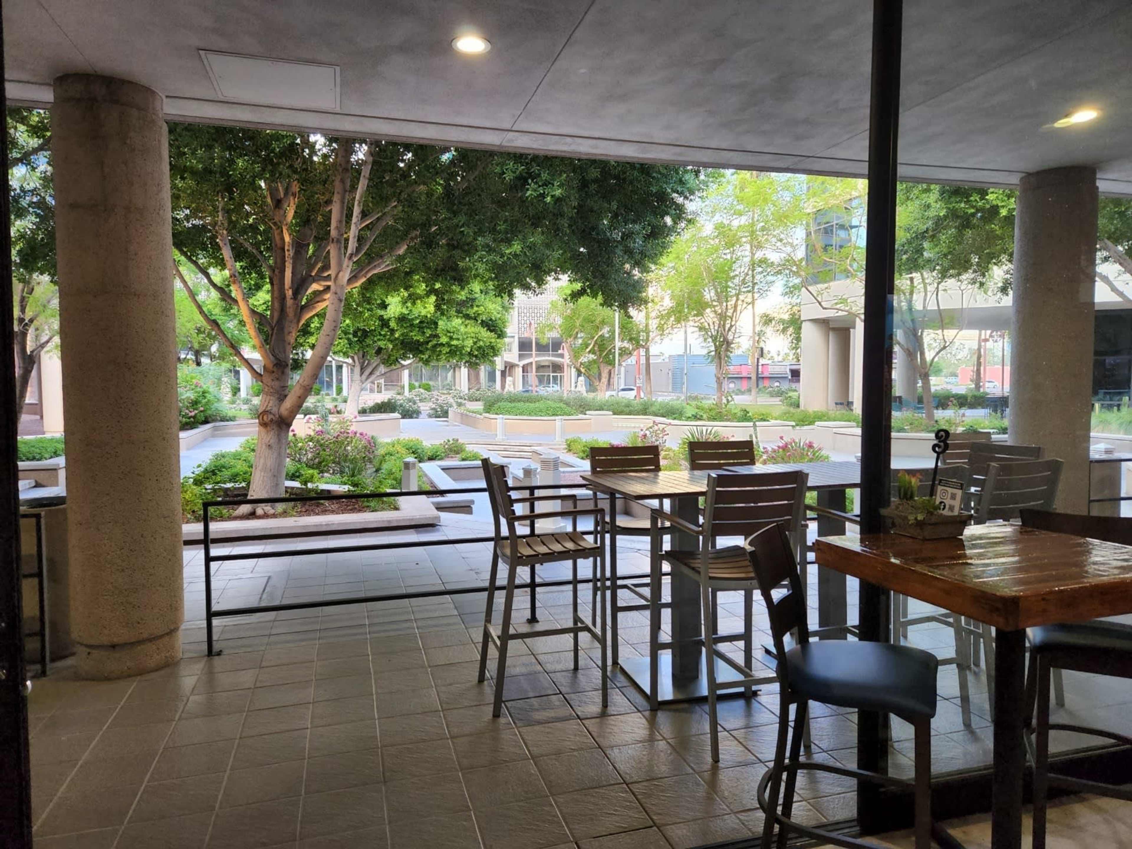 A view from inside a café, featuring tables and chairs arranged near large windows overlooking a landscaped outdoor area with trees and pathways.