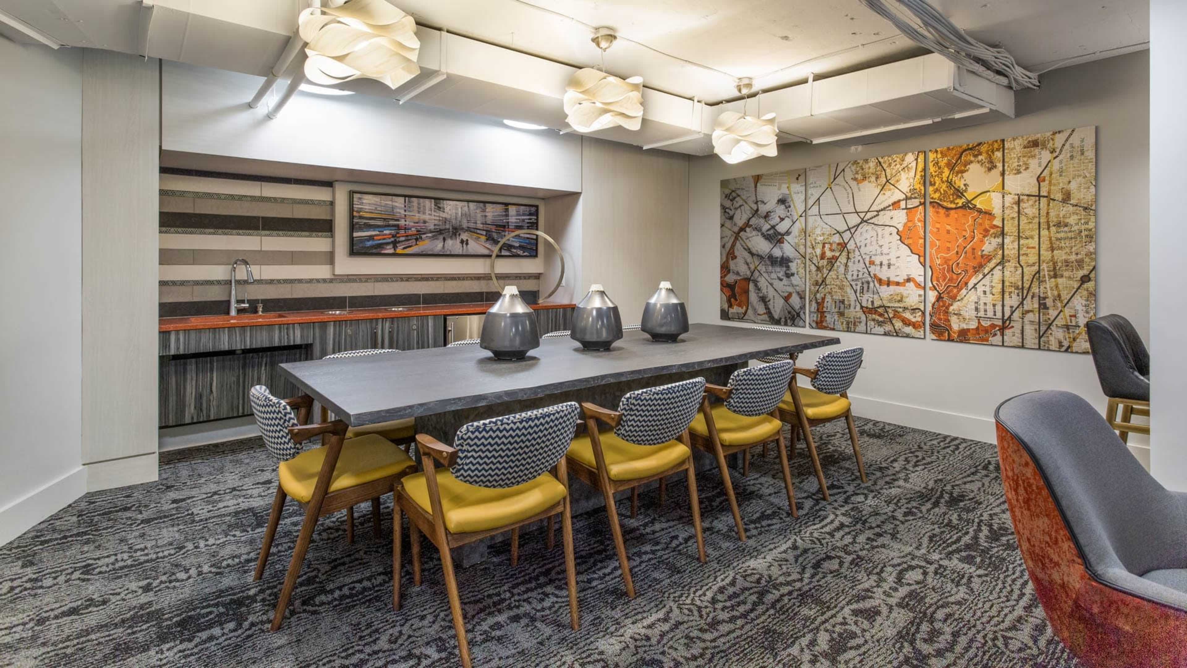A dining room features a long table with eight chairs, a wall-mounted artwork depicting a map, and a kitchenette area with a sink.