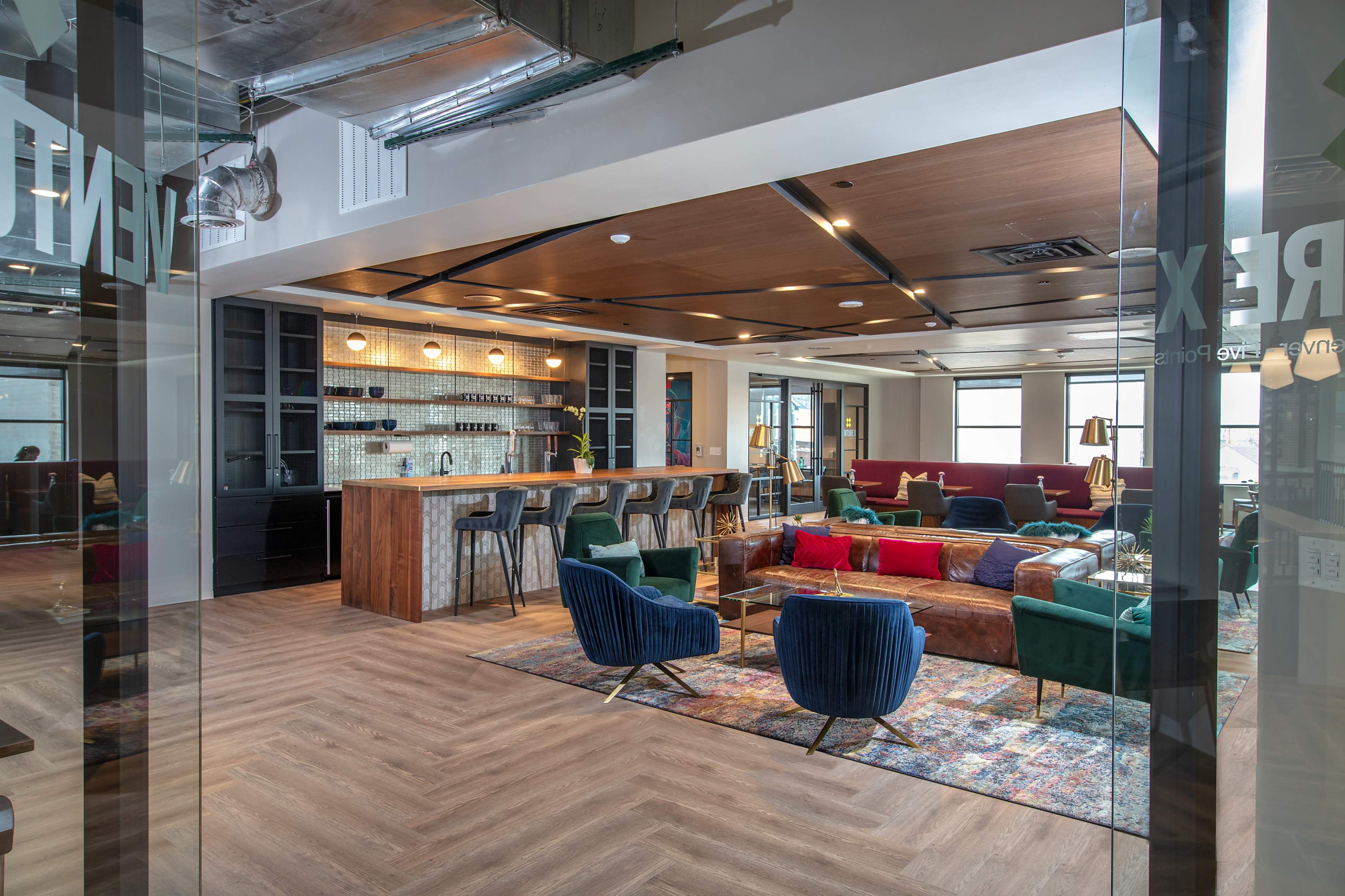 The image shows a modern office lounge area featuring a bar, seating arrangements with colorful furniture, and an open layout with wooden accents.