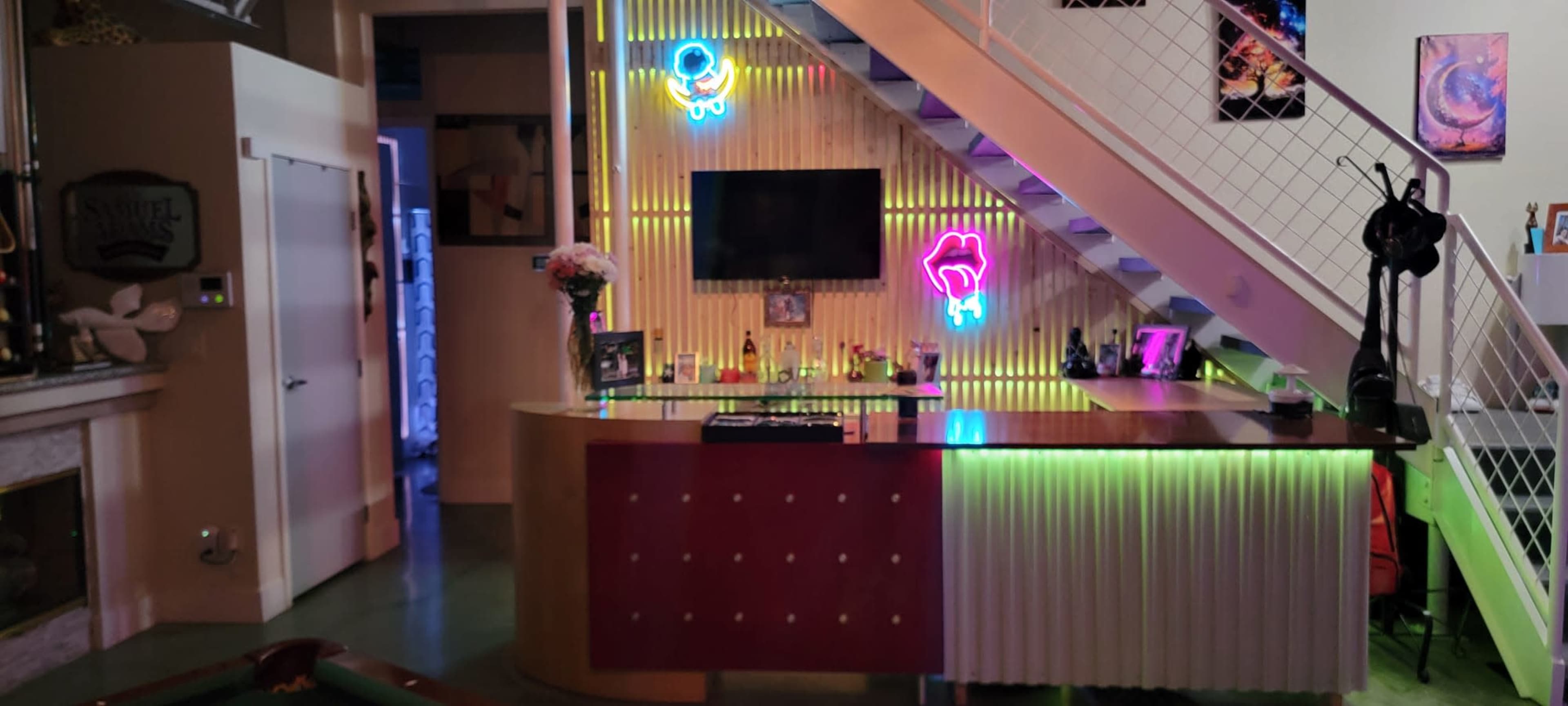 The image shows a bar area with colorful neon lights and a television, located in a modern, open space beneath a staircase.