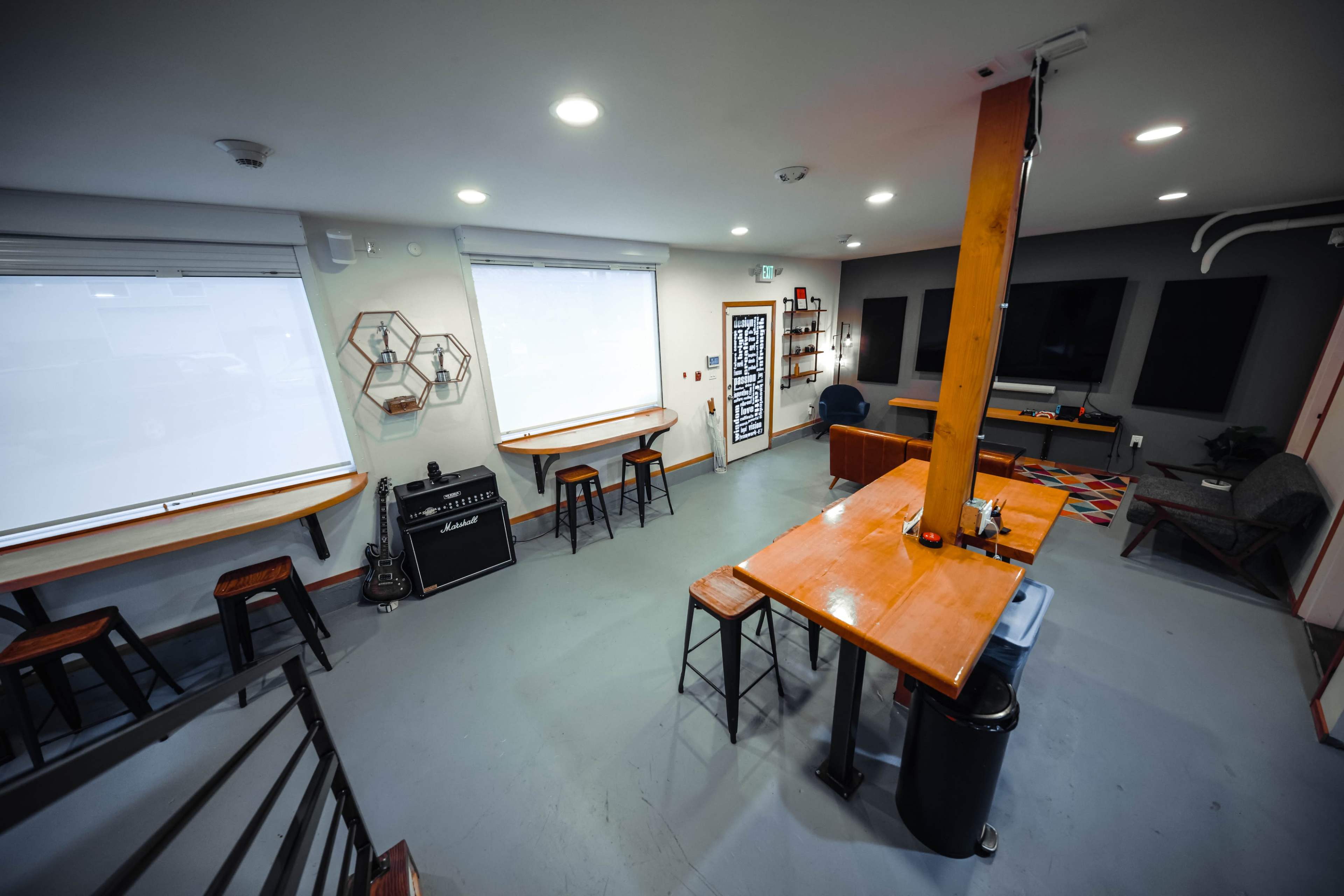 A modern common area with a wooden table, high stools, a guitar amplifier, and two large windows.