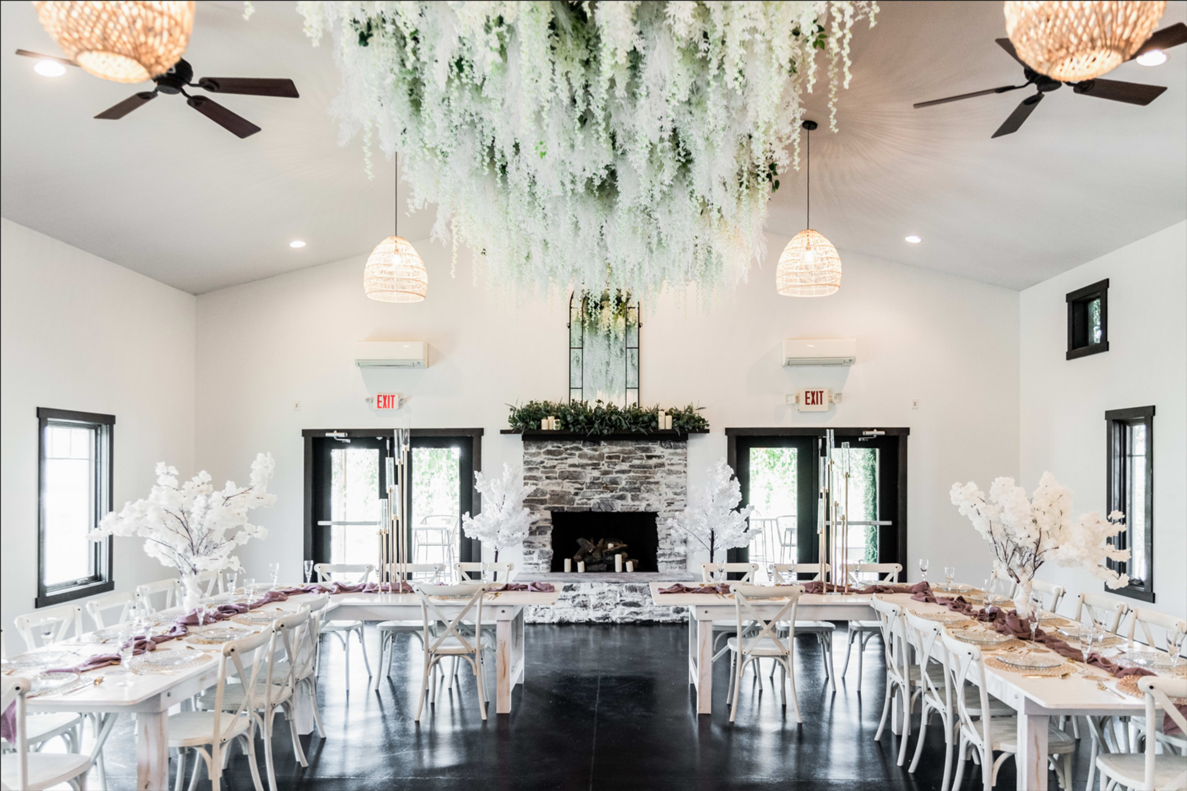 A spacious indoor venue features long dining tables set for an event, with floral decor hanging from the ceiling and white trees positioned along the tables.