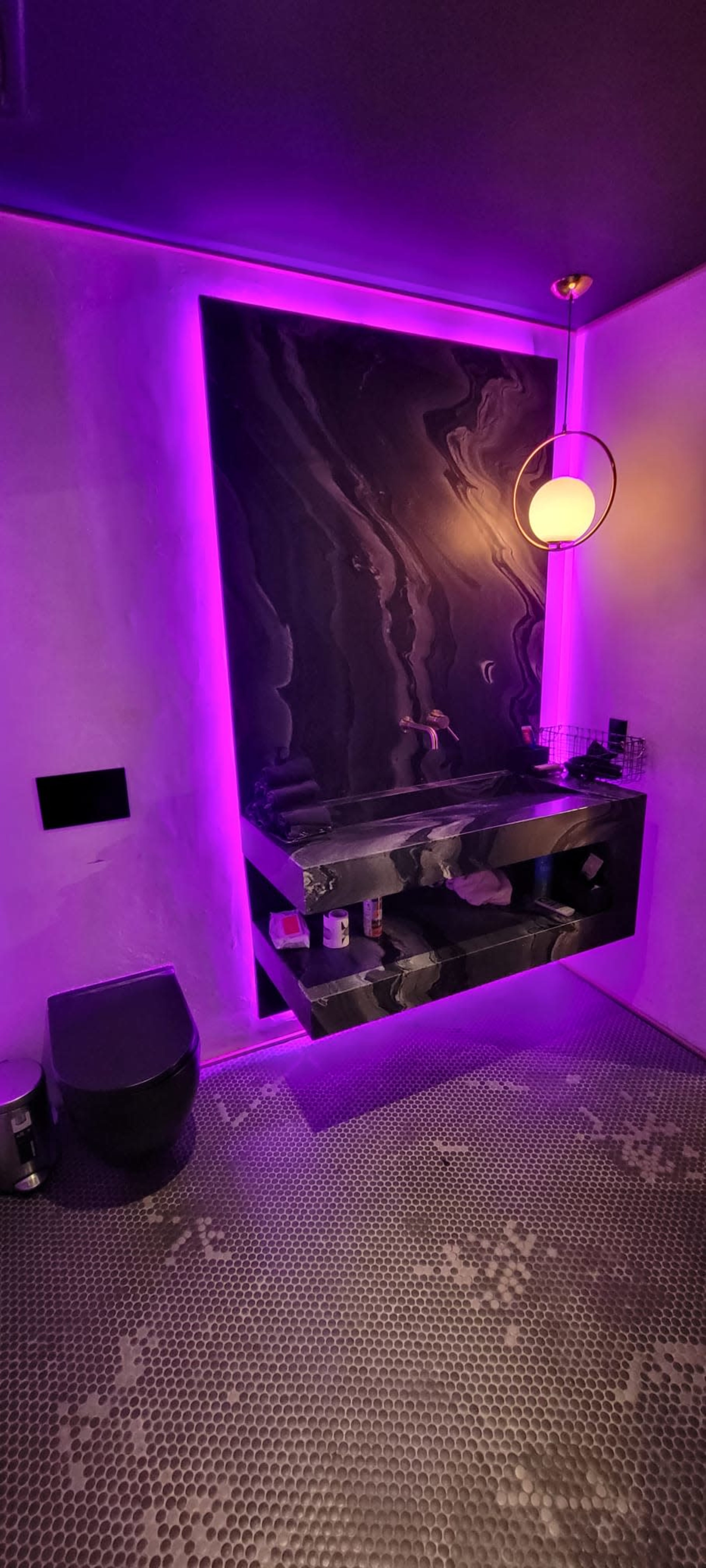 The image shows a modern bathroom featuring a dark marble vanity, a round light fixture, and purple ambient lighting.