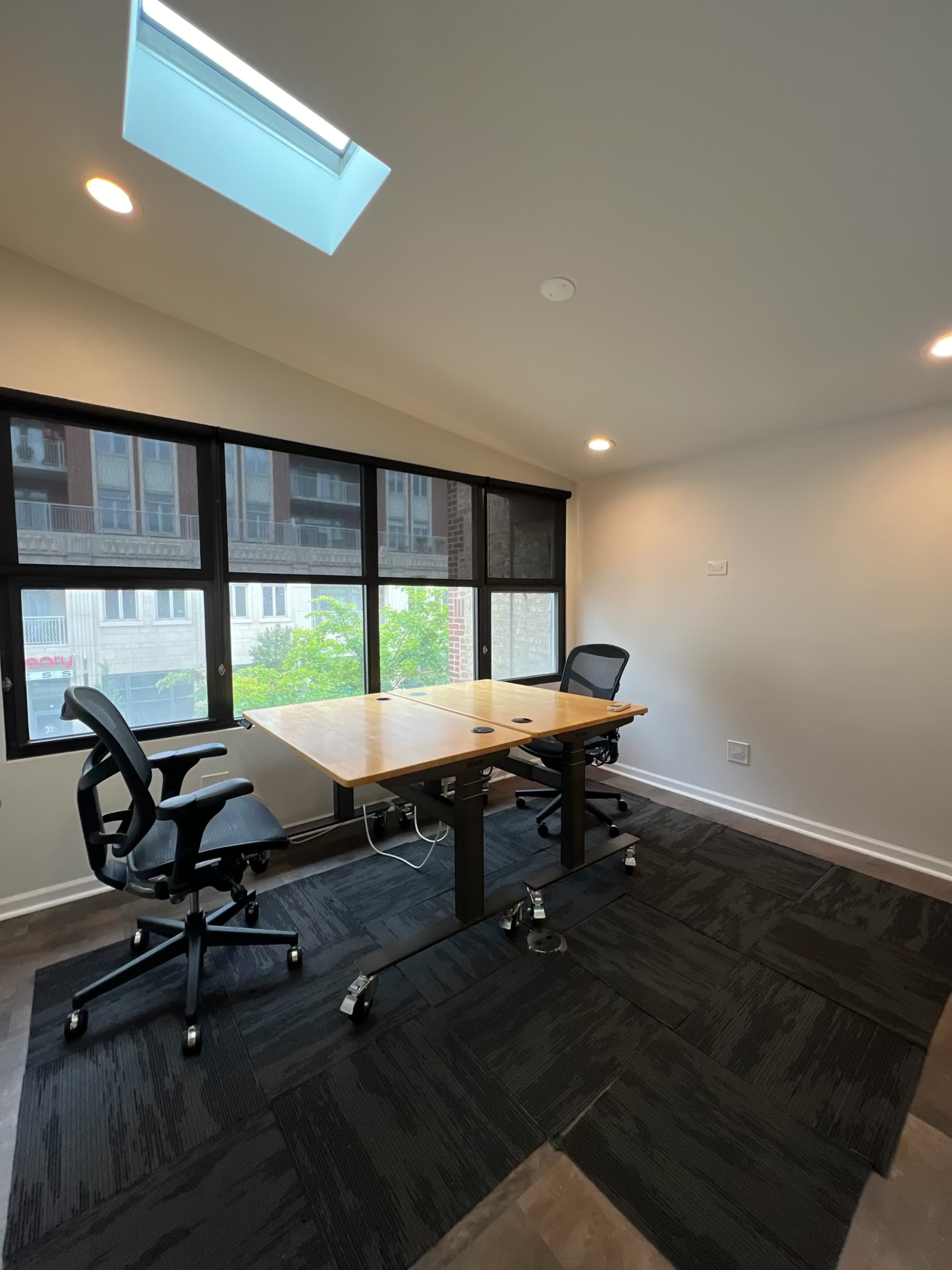 A small meeting room features a wooden table surrounded by two office chairs and large windows providing natural light.