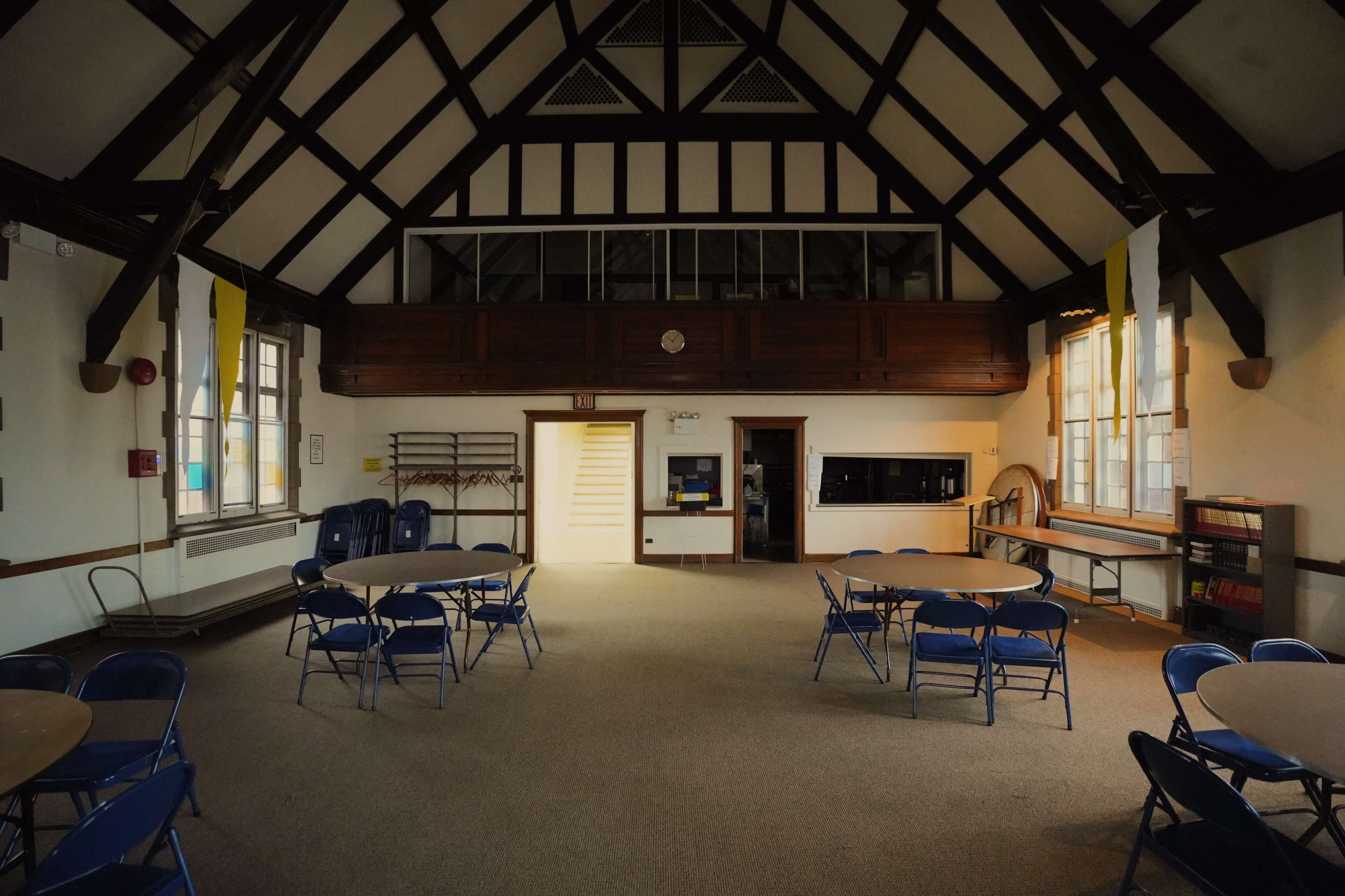 The image shows the interior of a large room with high, wooden-beamed ceilings, multiple blue folding chairs arranged around circular tables, and a kitchenette area in the background.
