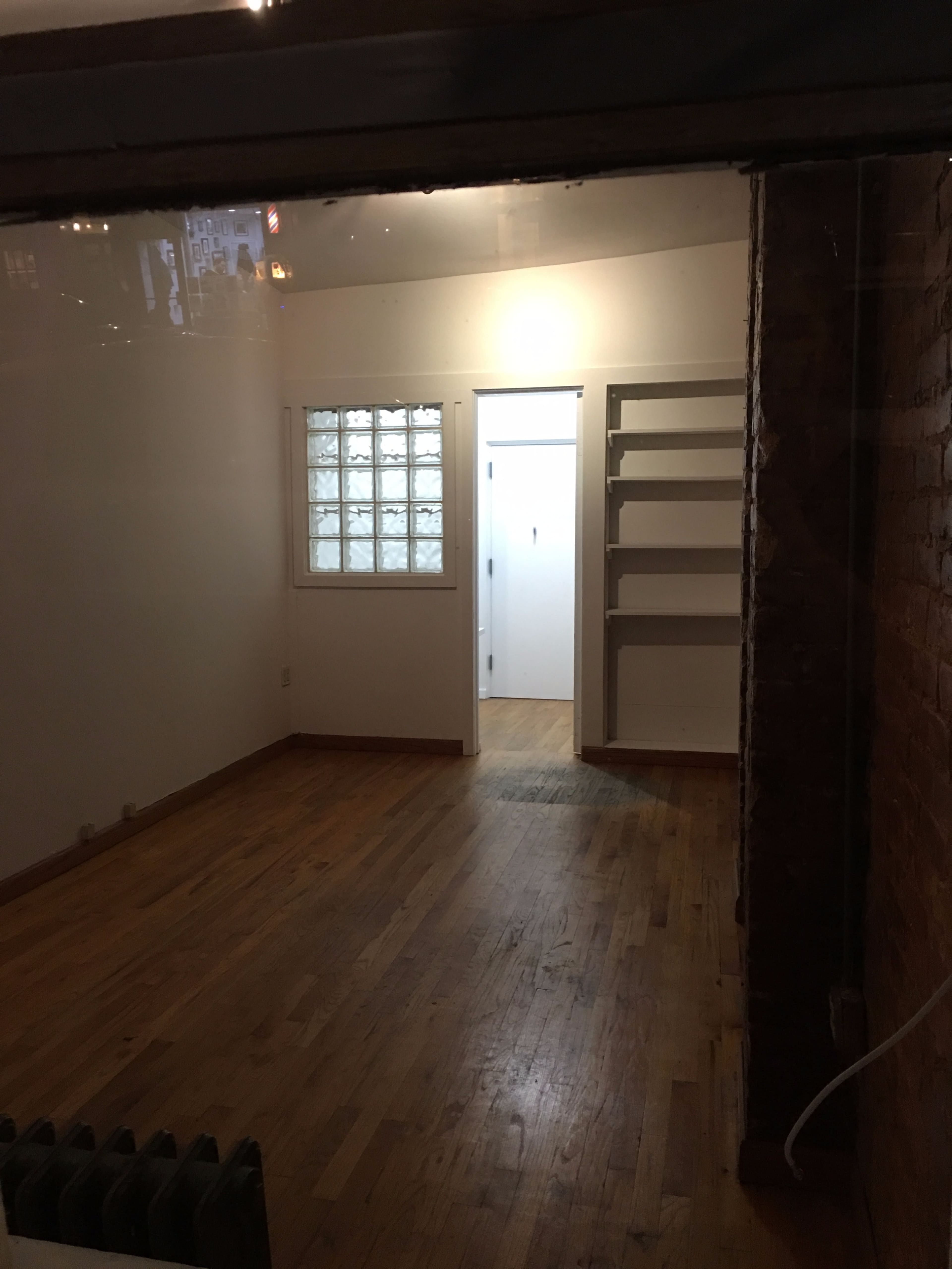 The image shows a sparsely furnished, empty room with wooden flooring, featuring a glass block window and an adjoining door leading to another space.