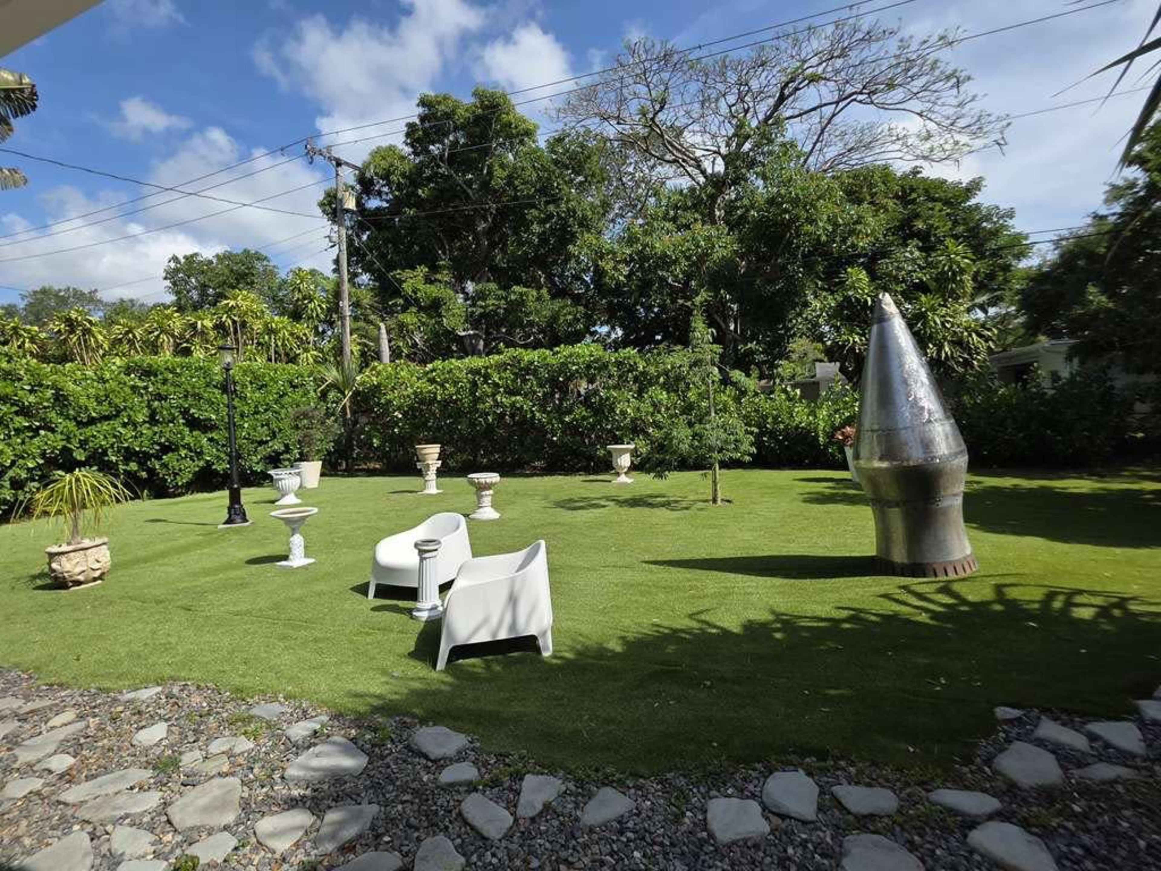 The scene features a landscaped garden with manicured grass, various sculptures, and white furniture under a clear blue sky.