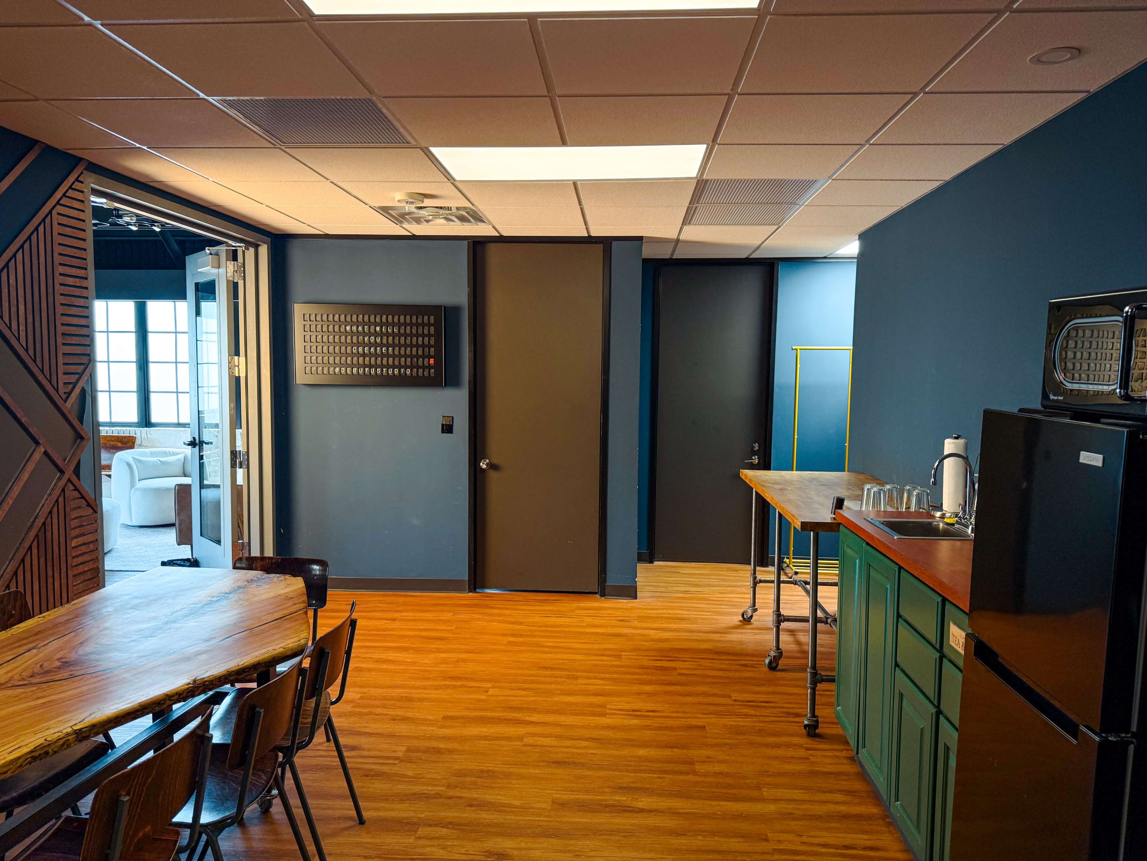 The image shows a spacious room with a wooden dining table, green cabinetry, a black refrigerator, and two doors leading to other areas.
