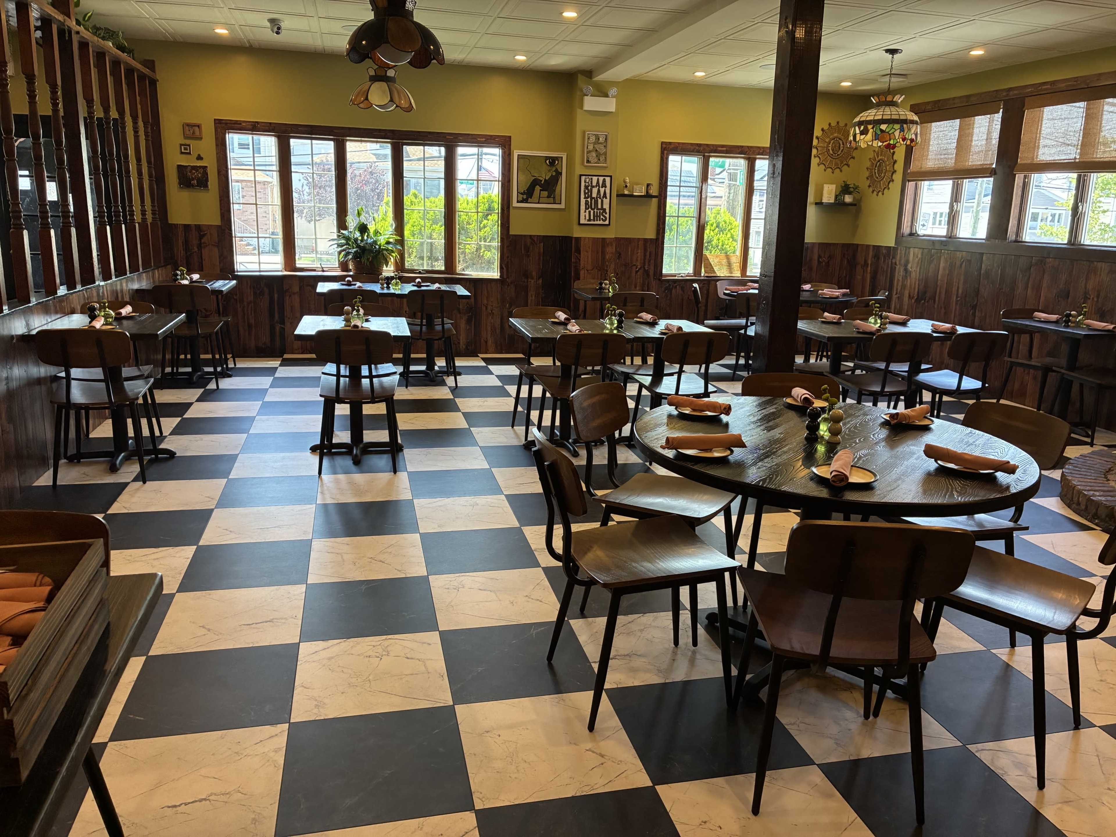 The image shows a restaurant interior featuring wooden tables and chairs arranged on a black and white checkered floor, with plants and decor on the walls.