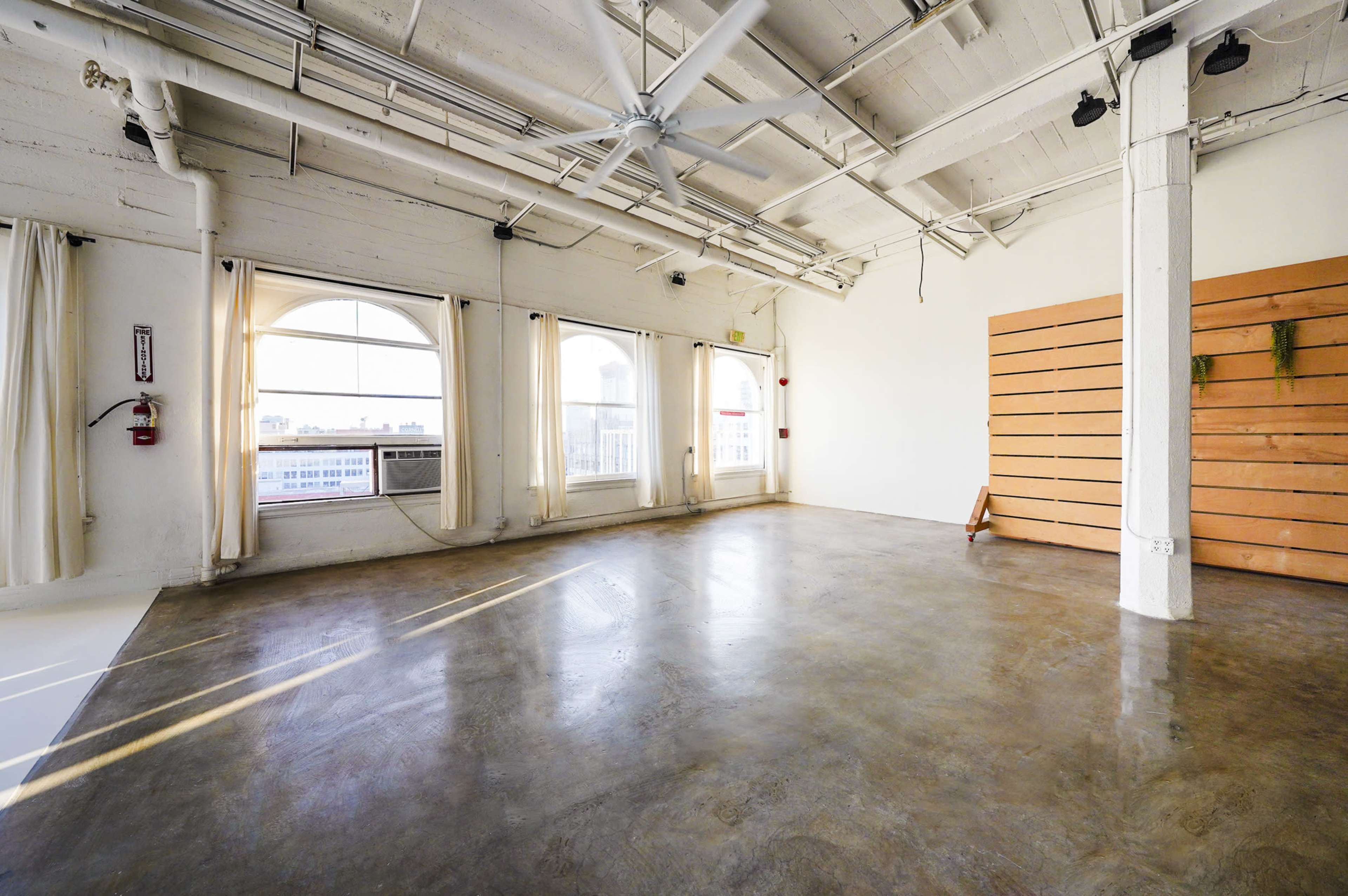 The image shows an empty, sunlit loft space with large windows, polished concrete floors, and wooden wall accents.