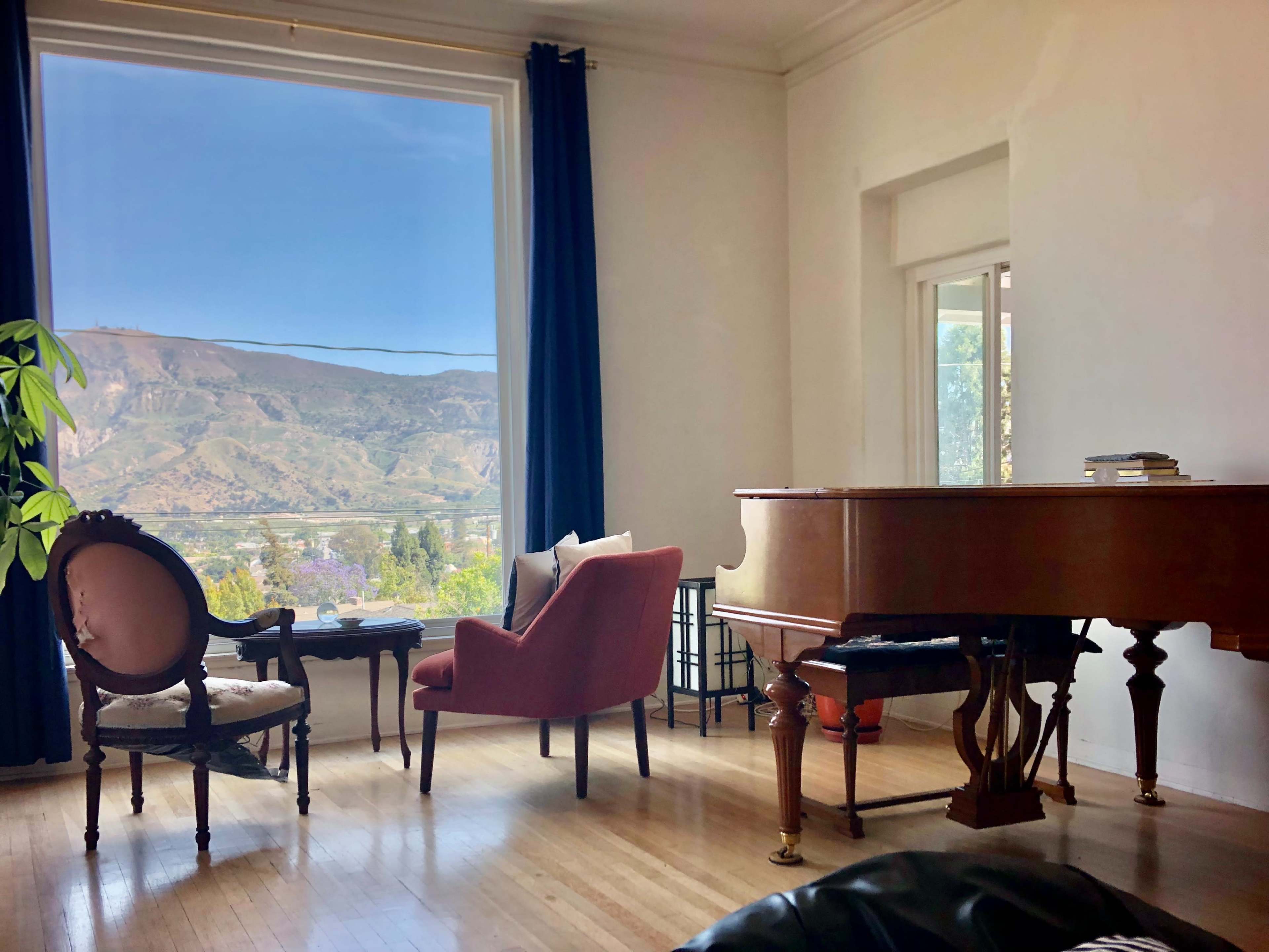 A grand piano and an armchair are positioned in a room with large windows overlooking a mountainous landscape.