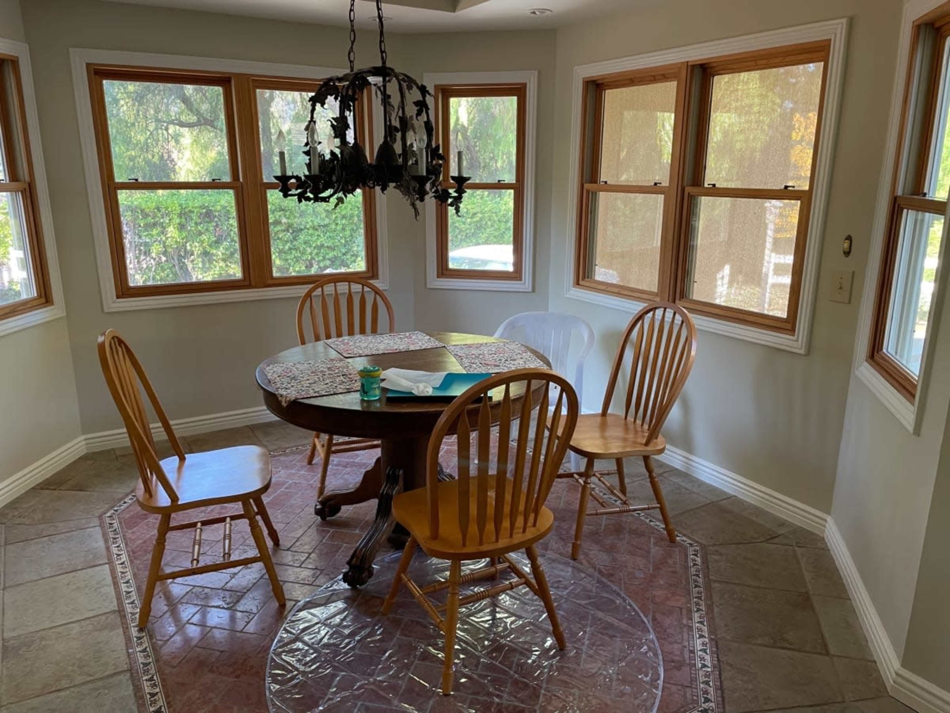 A round wooden table surrounded by four chairs is positioned in a sunlit dining area with large windows.