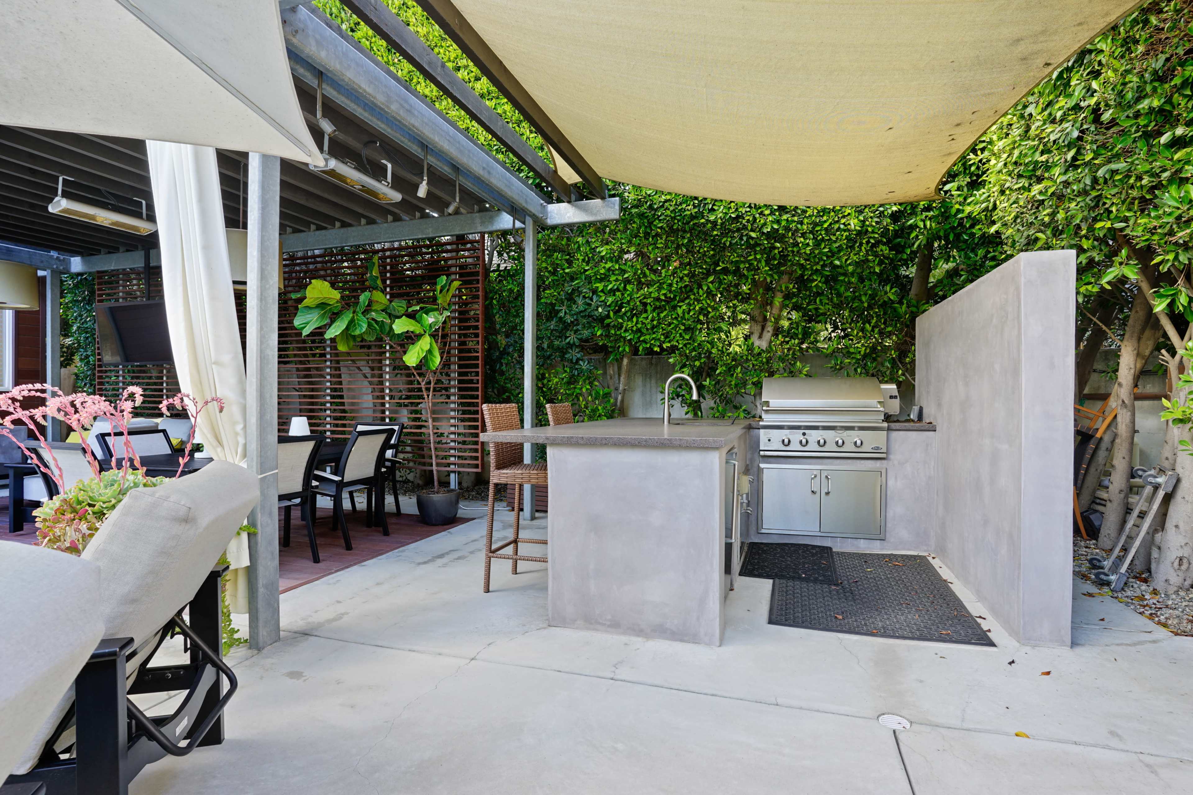 The image shows an outdoor kitchen area featuring a built-in grill, a counter, bar stools, and a shaded dining space surrounded by greenery.