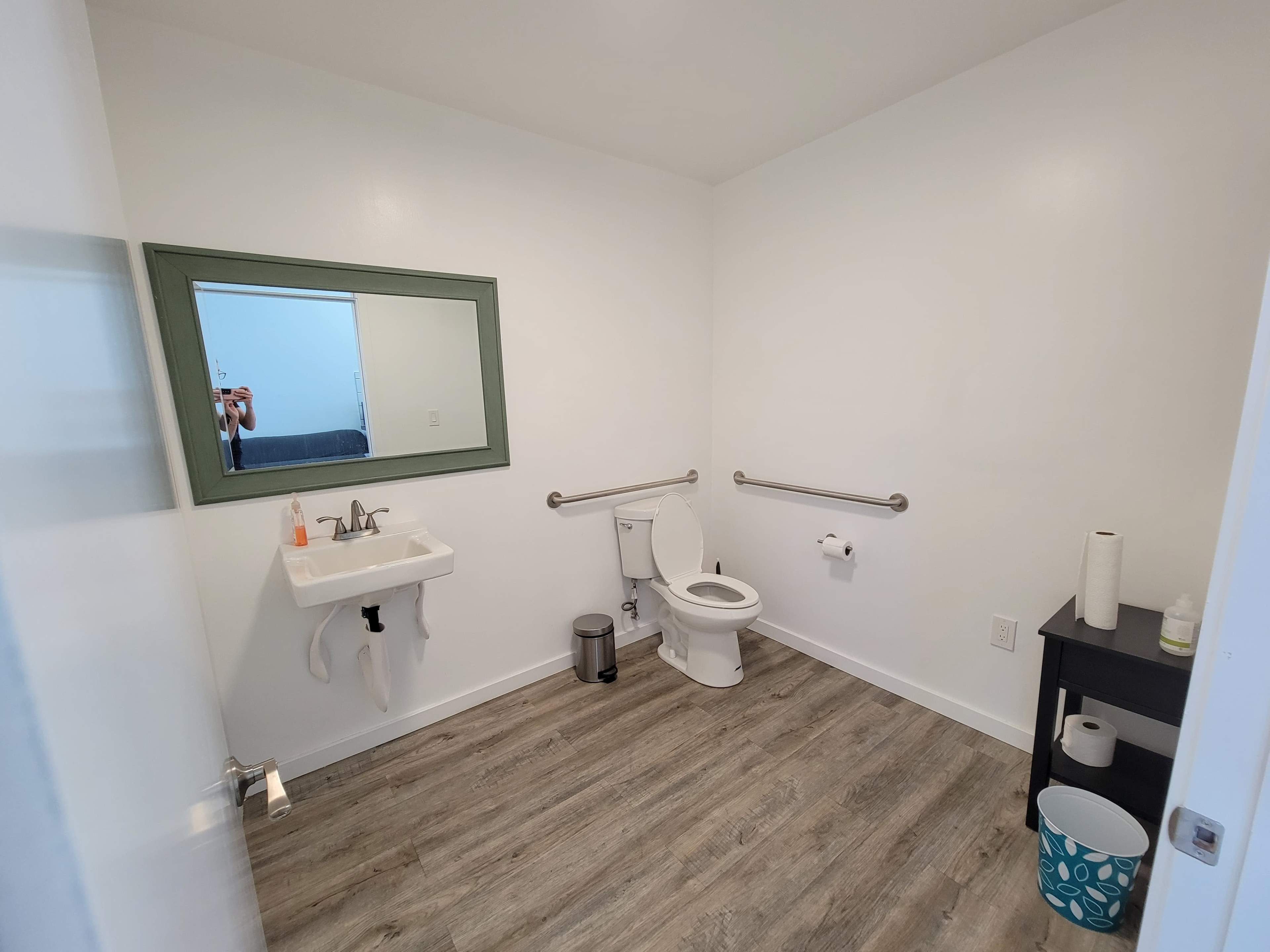 The image shows a clean, modern bathroom with a sink, toilet, and a large wall mirror, featuring grab bars and a simple storage table.