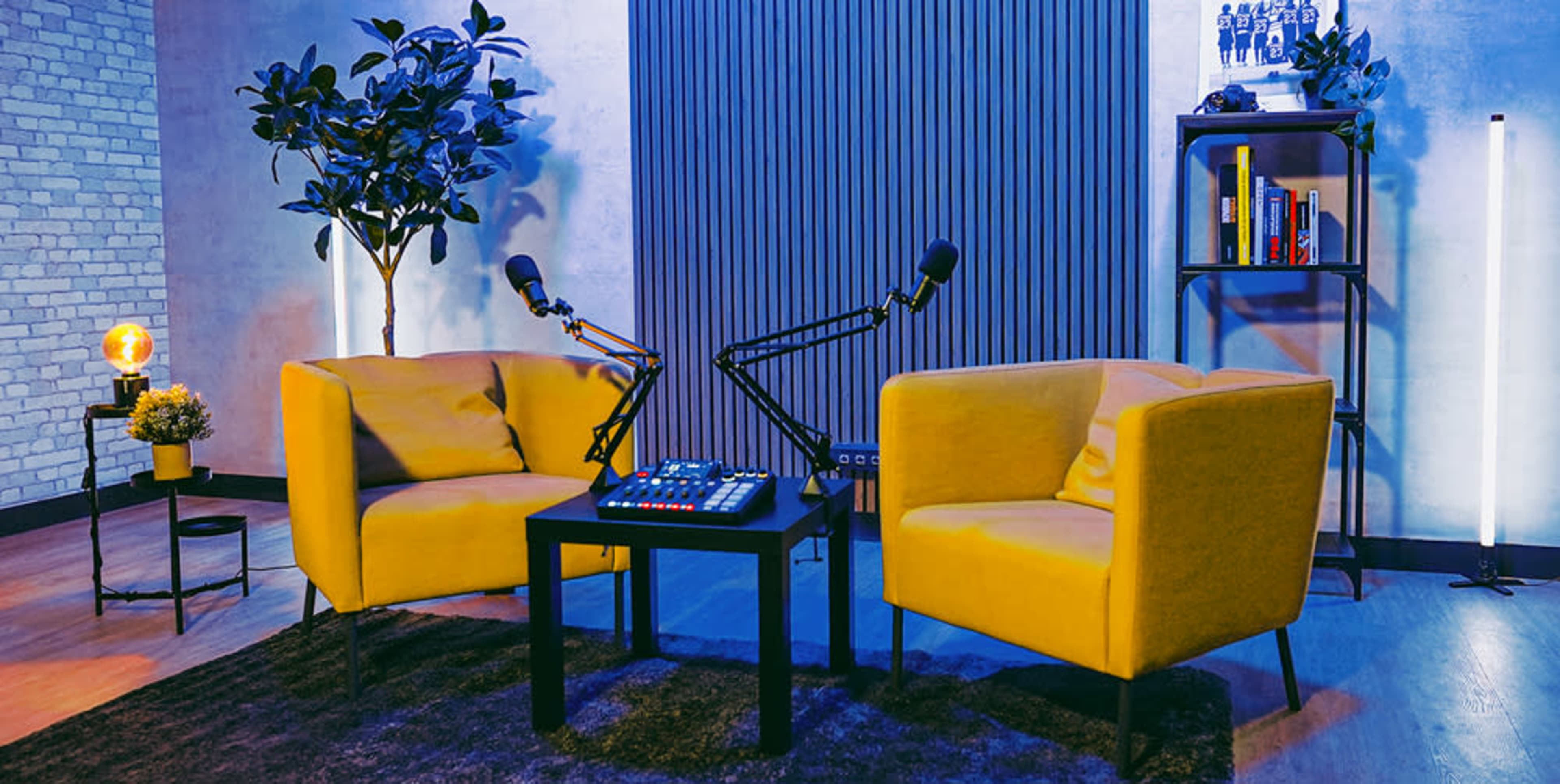 The scene features two yellow armchairs facing each other with a black table in between, surrounded by decorative plants and lighting in a modern studio environment.
