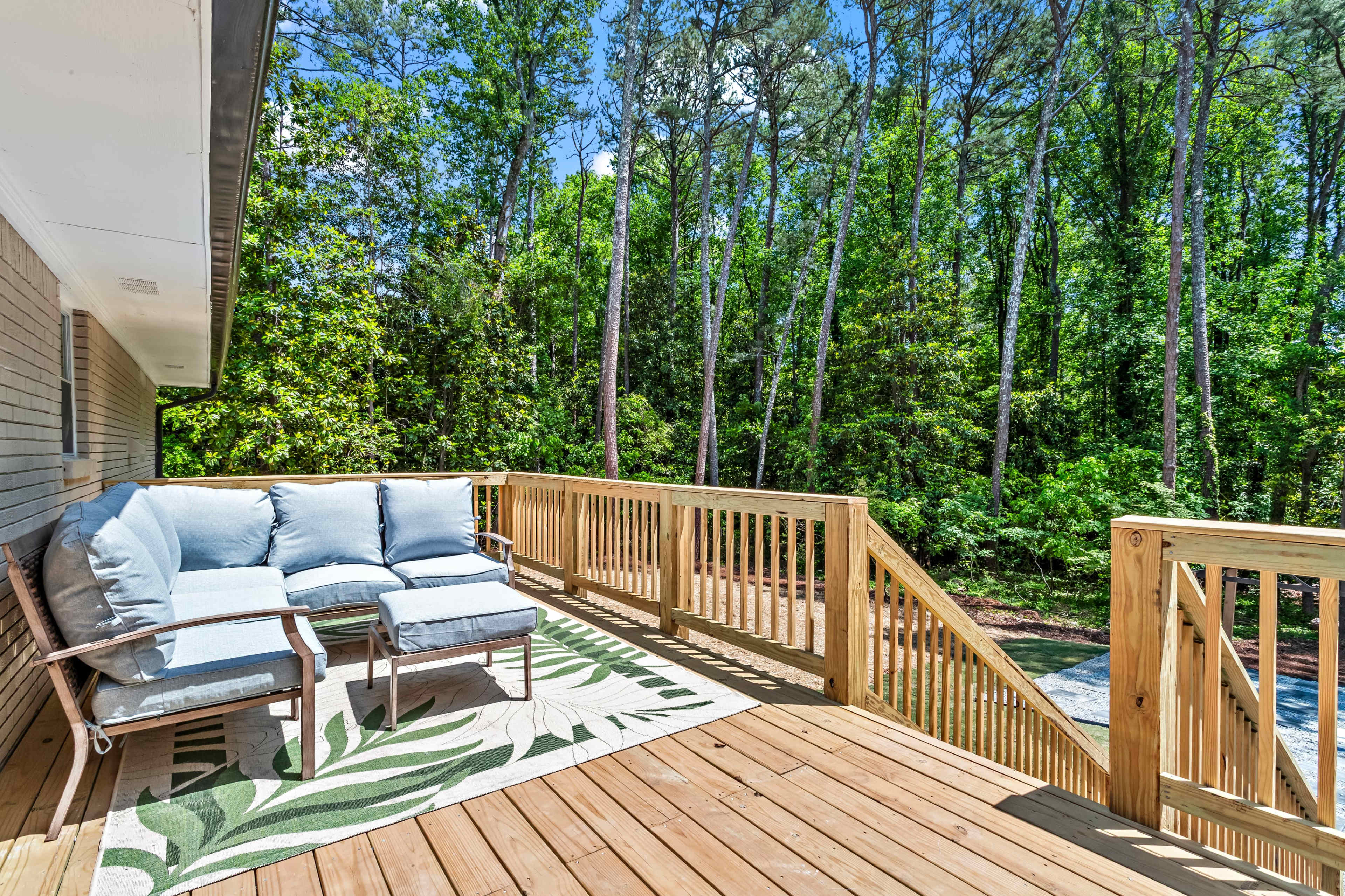 A wooden deck features a seated area with blue cushions and a grassy outdoor rug, surrounded by tall trees.