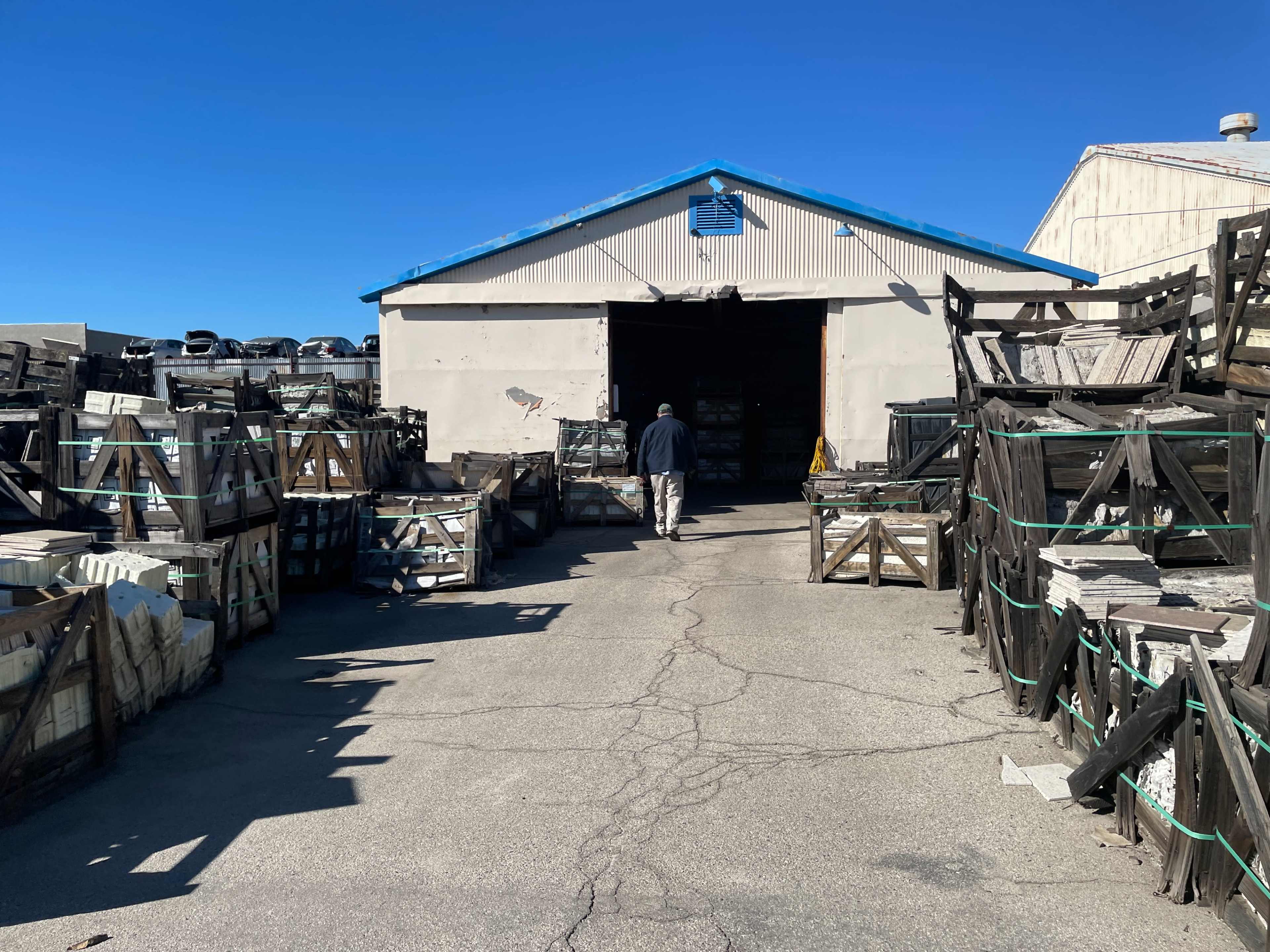 A person walks towards a large storage building surrounded by stacked crates and containers under a clear blue sky.
