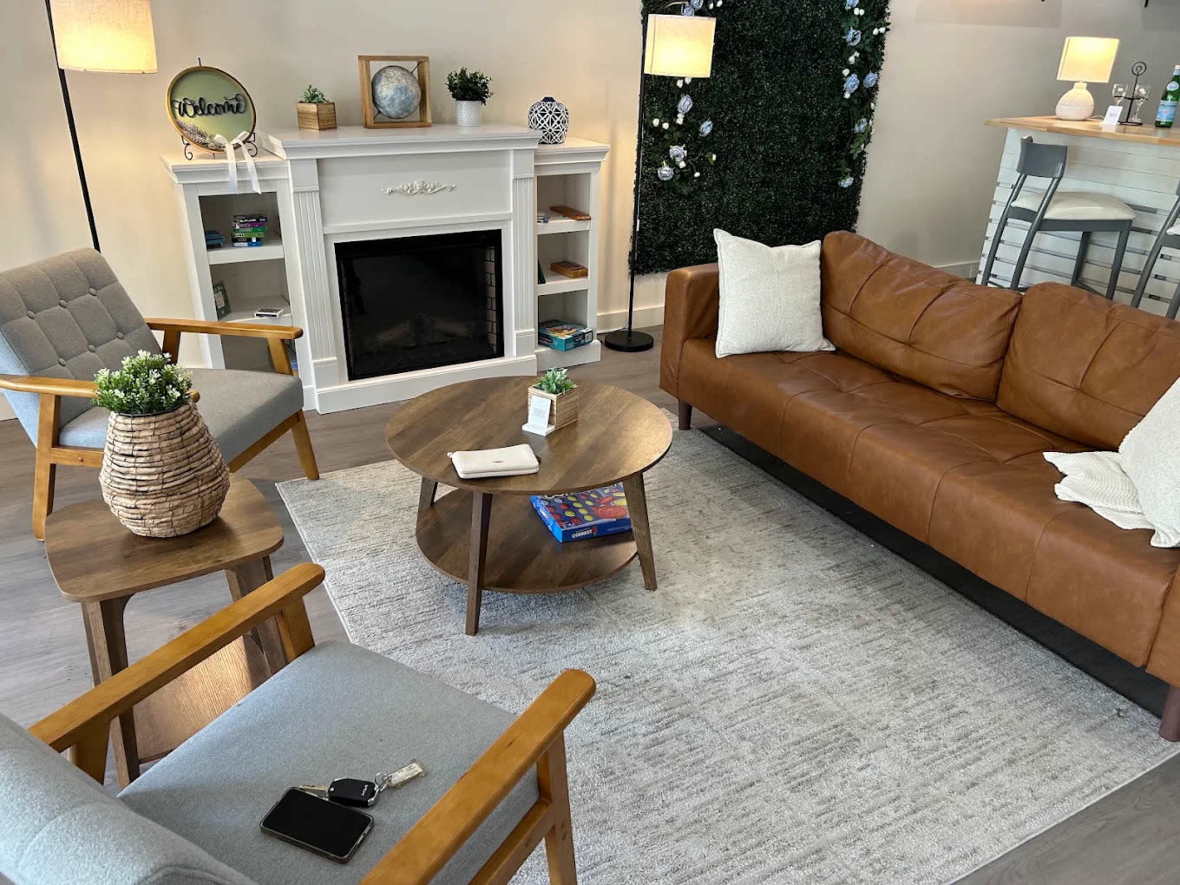 The image shows a cozy living room with a brown leather sofa, two wooden chairs, a round coffee table, and a decorative fireplace.