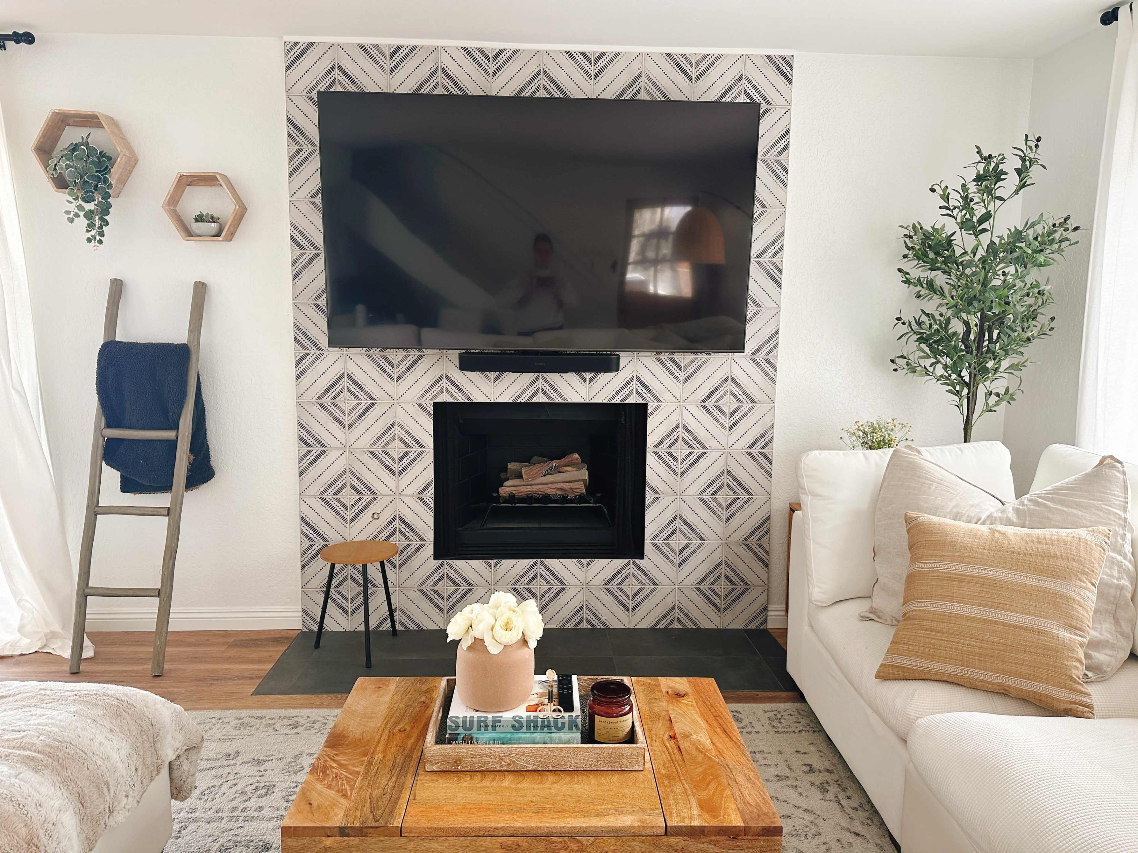 A modern living room features a patterned accent wall with a large flat-screen TV above a black fireplace, surrounded by a white sofa, a wooden coffee table, and decorative plants.