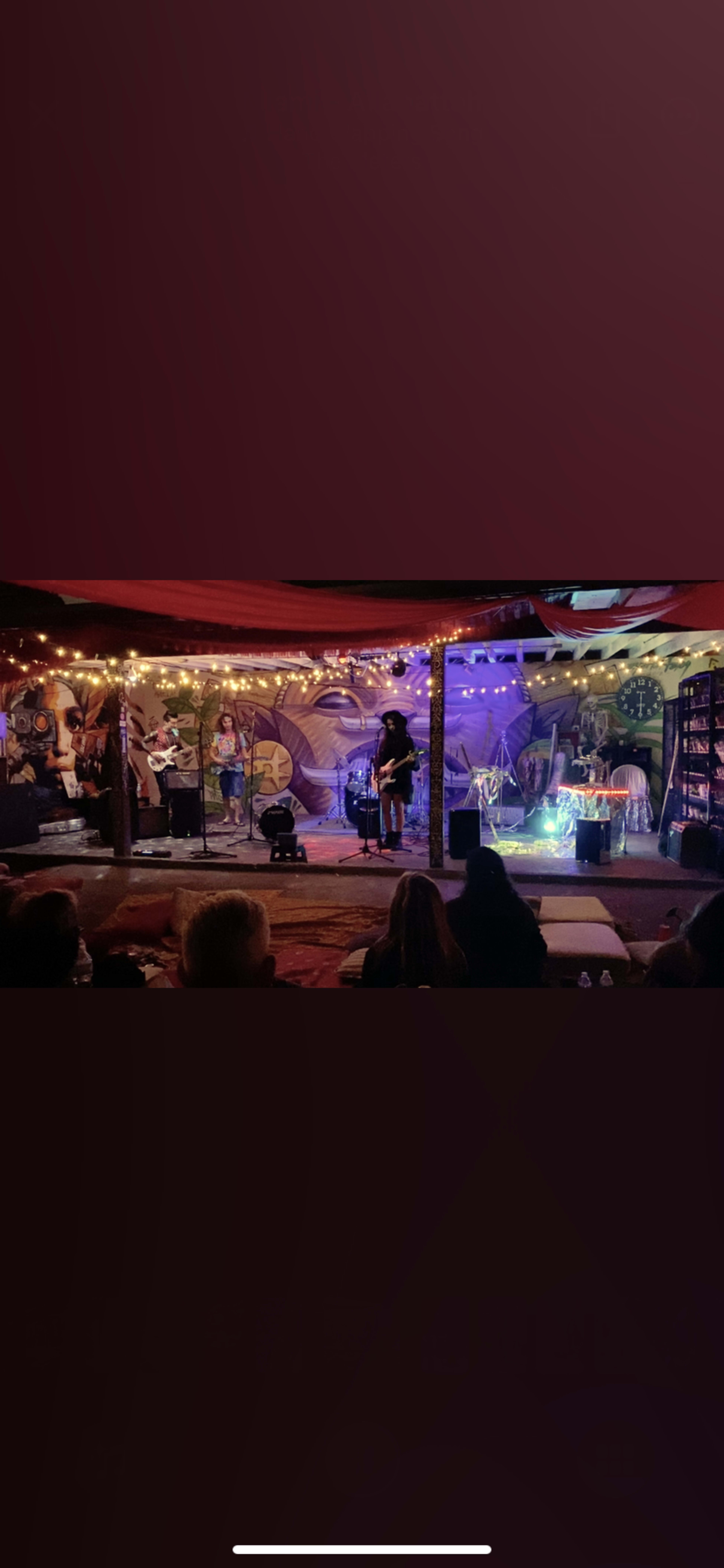 A small indoor stage is set up for a performance, adorned with colorful murals and string lights, while an audience watches from seats in the foreground.