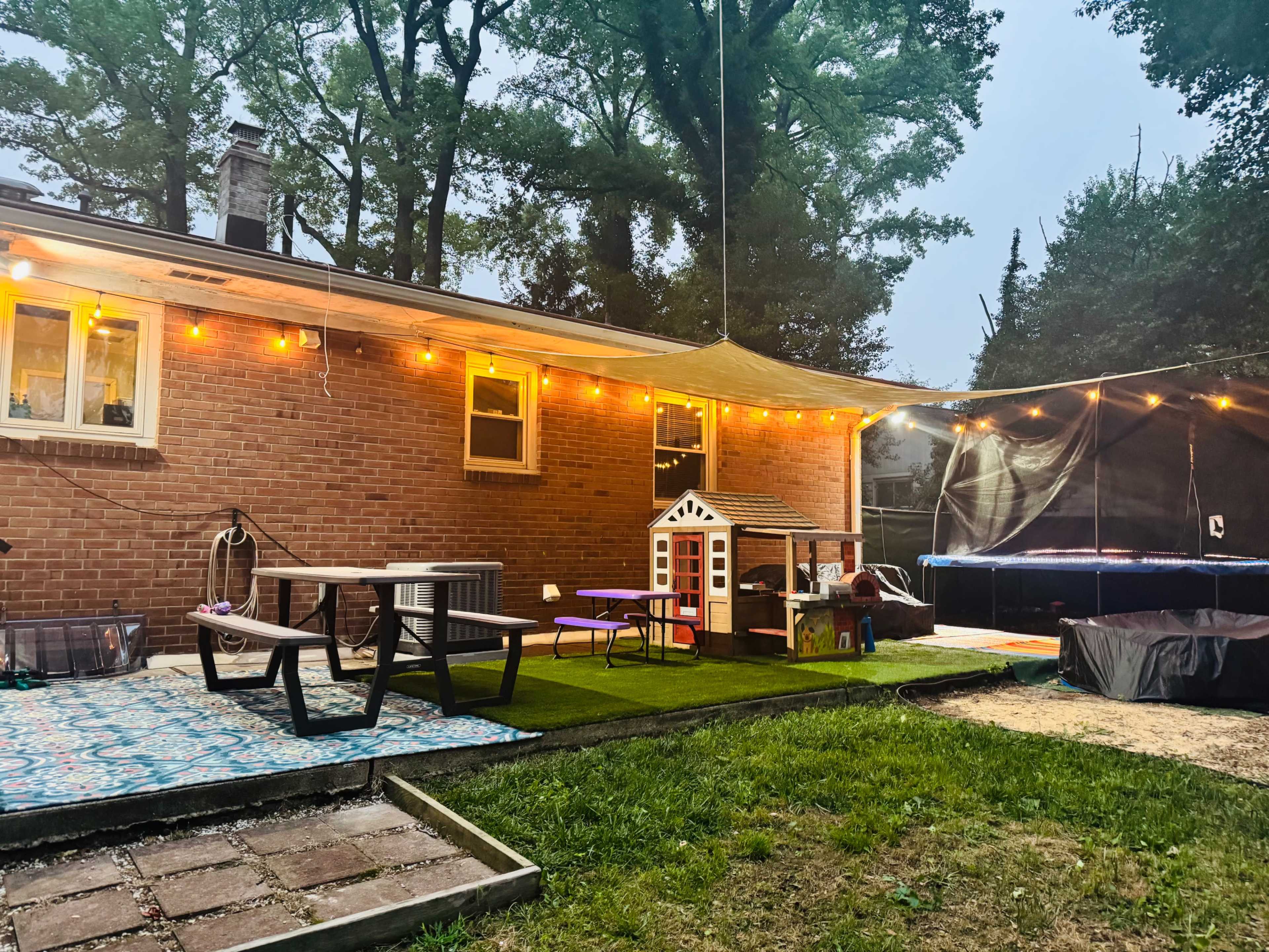 A backyard area features a brick house with string lights, a playhouse, a trampoline, and a picnic table on green grass.