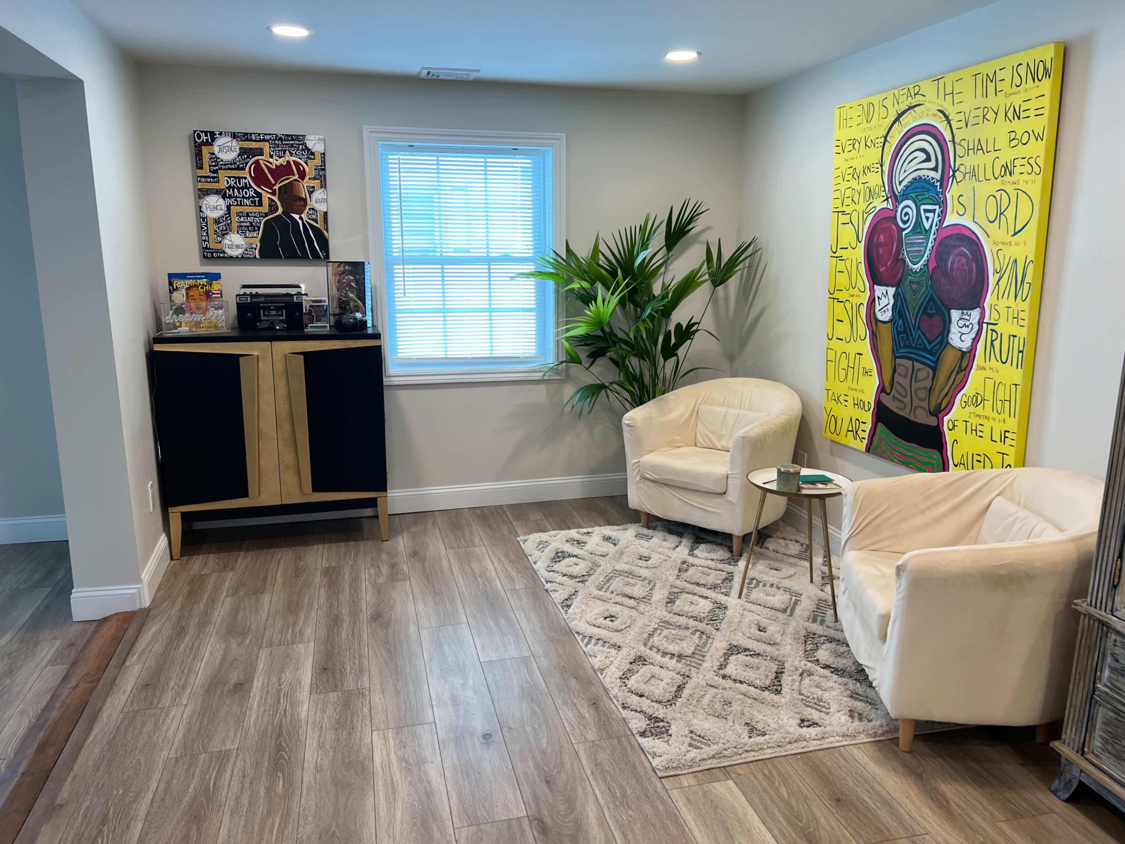 A stylish living space featuring two white chairs facing a small table, a patterned rug, a corner plant, and a large colorful artwork on the wall.