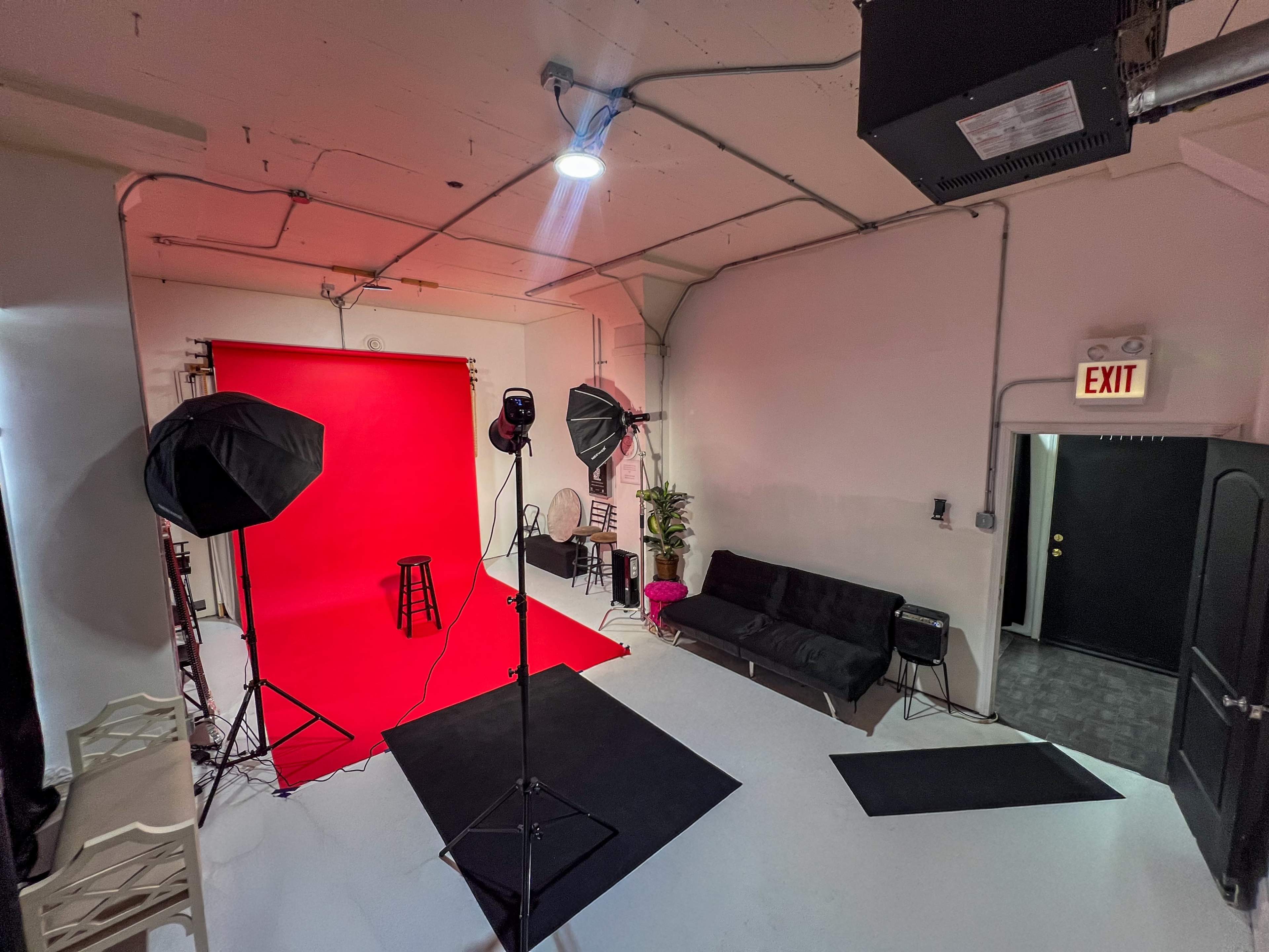 The image shows a photography studio featuring a red backdrop, lighting equipment, and a sofa in a well-lit space.
