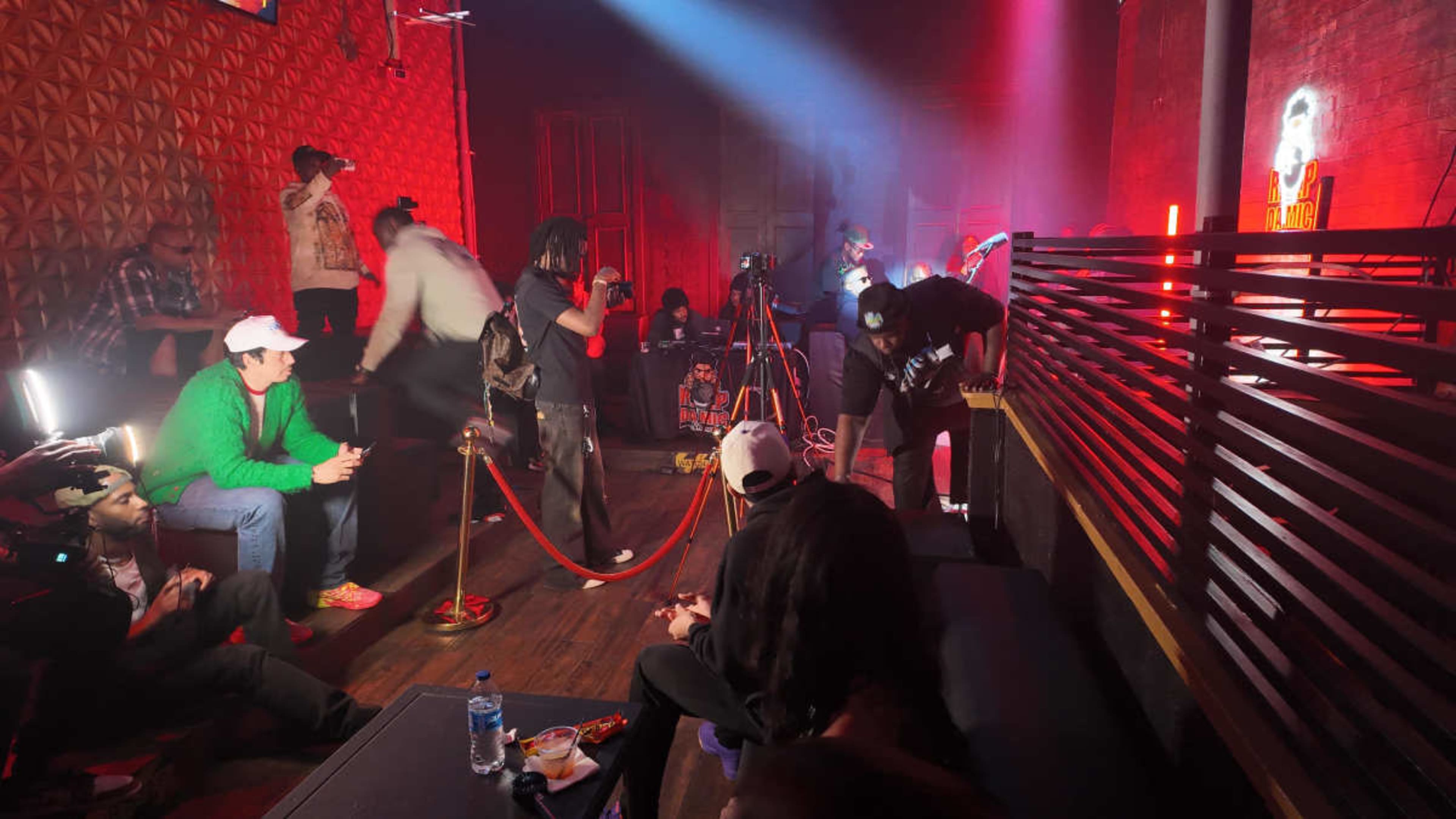 A group of people is gathered in a dimly lit venue, some setting up cameras and lighting, while others sit on benches and the floor.