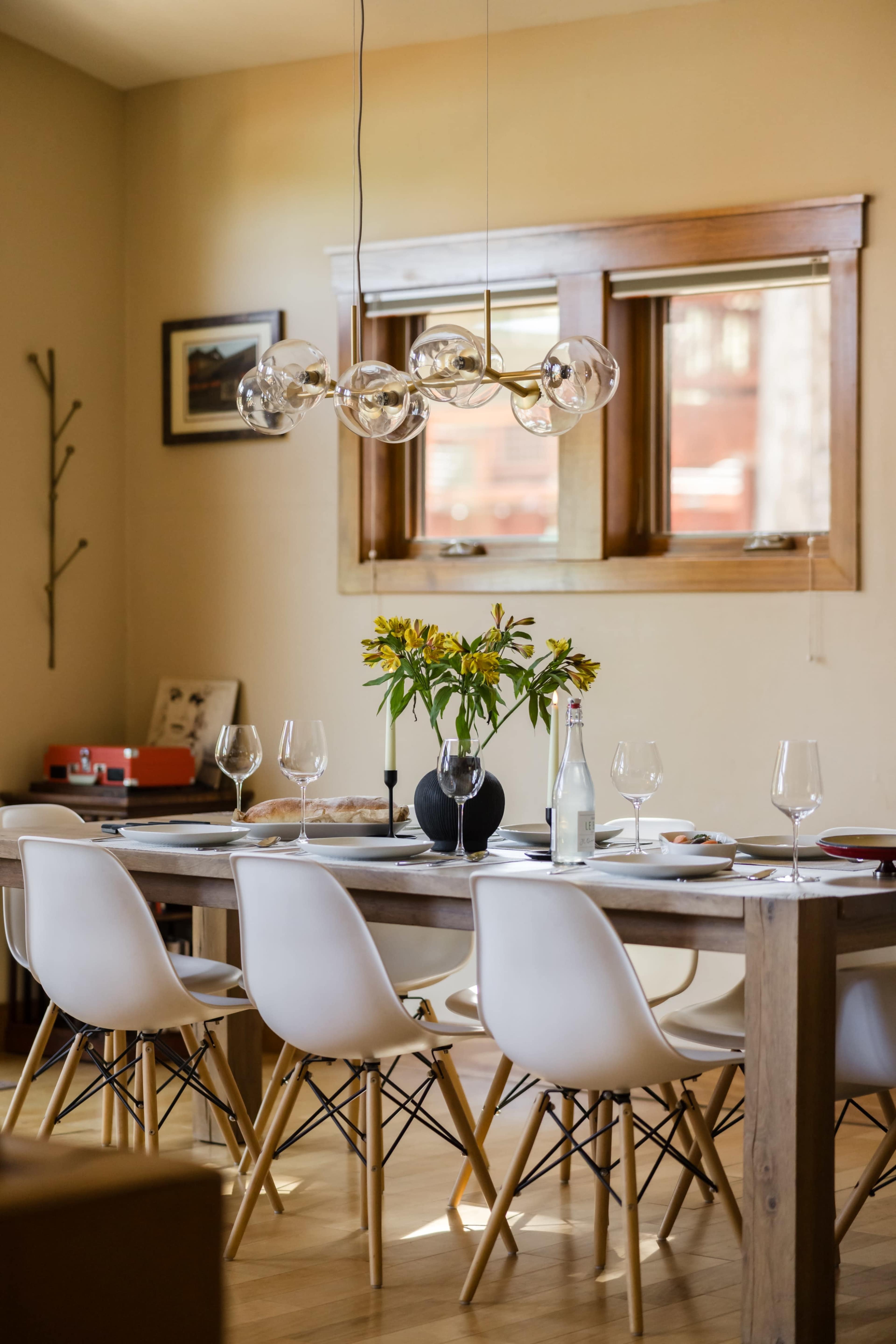 A long wooden dining table is set with glassware, a vase of flowers, and modern chairs, illuminated by a contemporary light fixture.