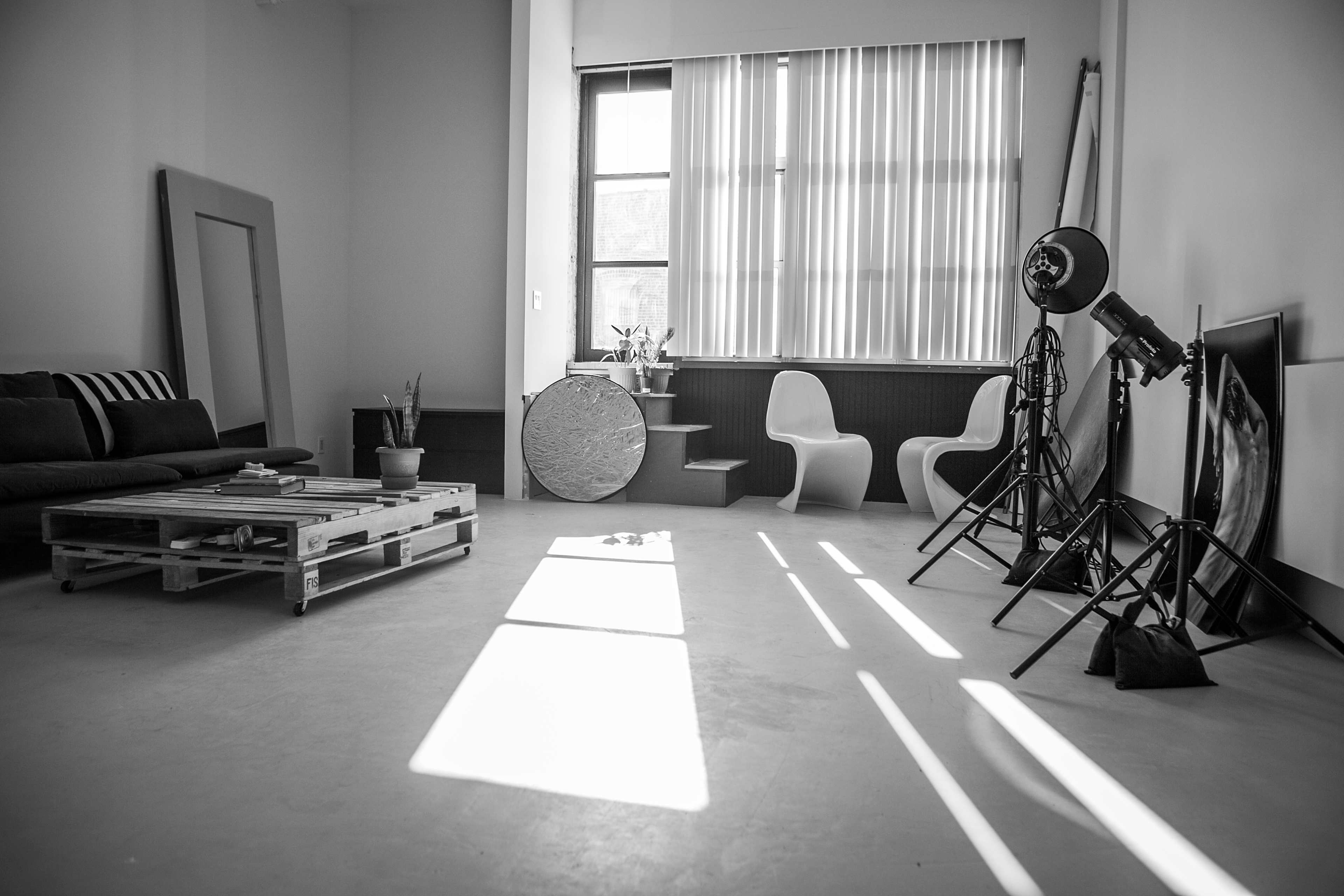 The image shows a minimalist interior space with a wooden pallet sofa, two modern chairs, a circular table, and photography lighting equipment.