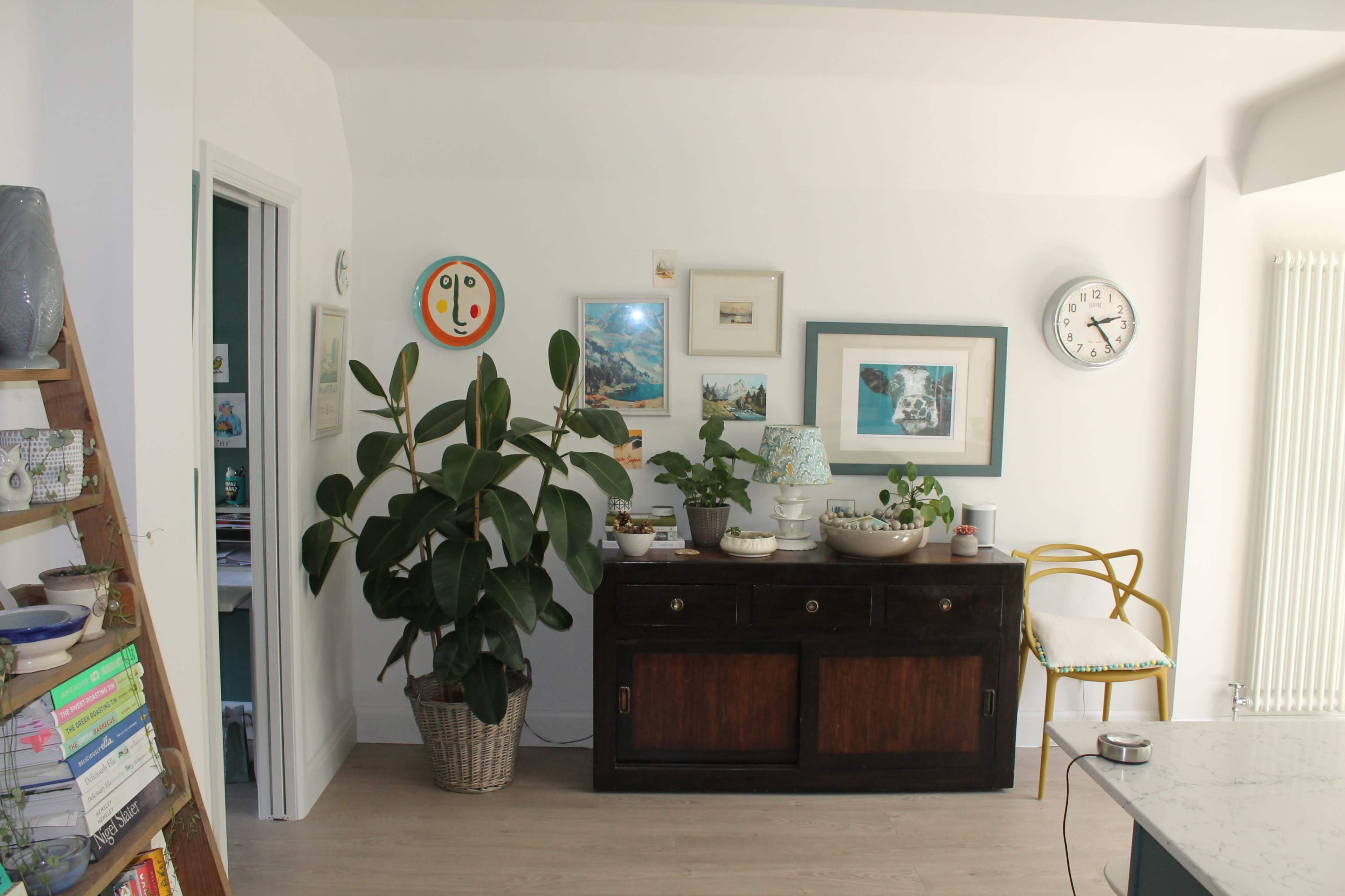 A bright room featuring a wooden sideboard adorned with plants and decorative items, with a wall displaying various artworks and a clock.