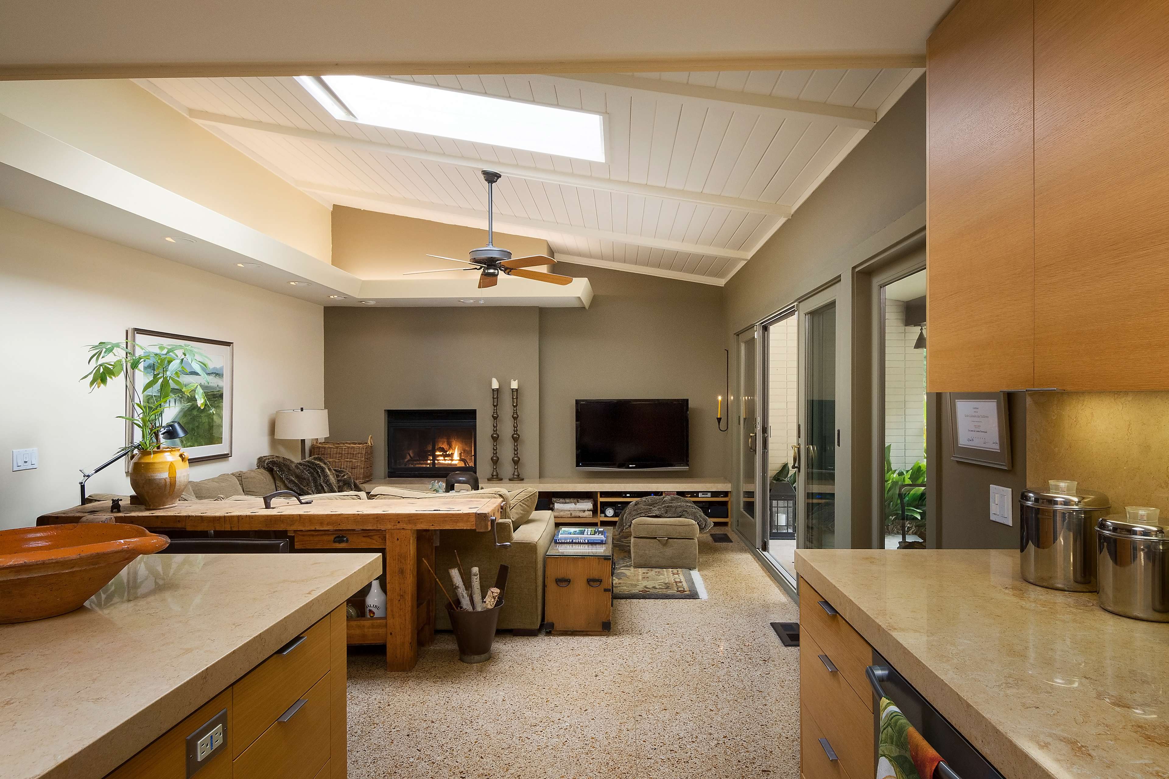 The scene depicts a modern living room with a fireplace, large windows, and an open kitchen area featuring wooden cabinetry and a countertop.